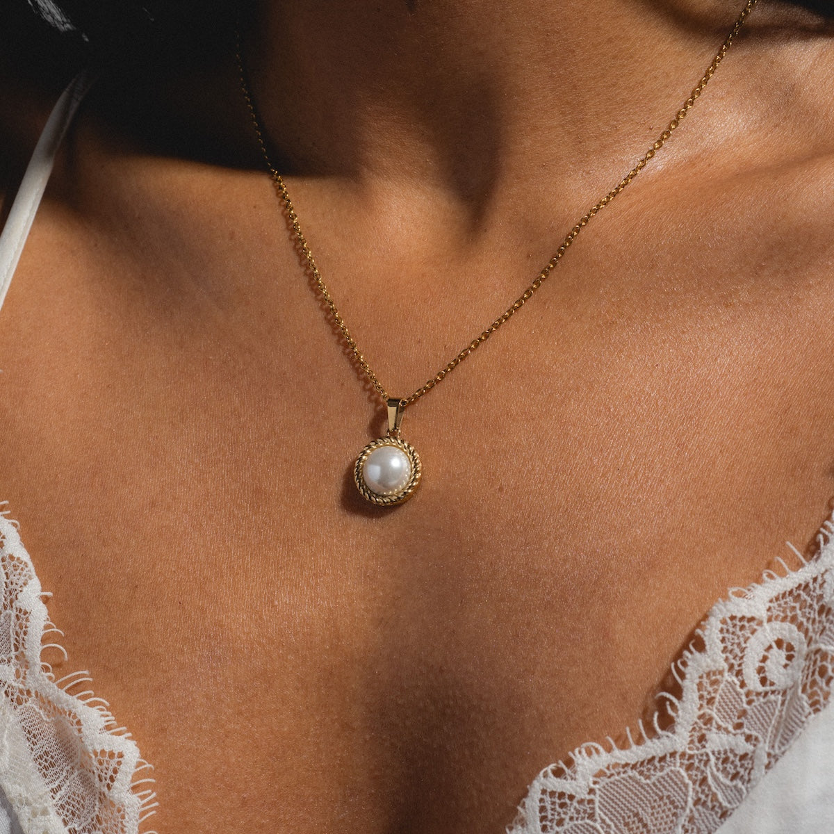 A close-up of a person wearing the Paloma Necklace—a yellow gold plated chain with a round freshwater pearl pendant—shown against bare skin and white lace-trimmed clothing.