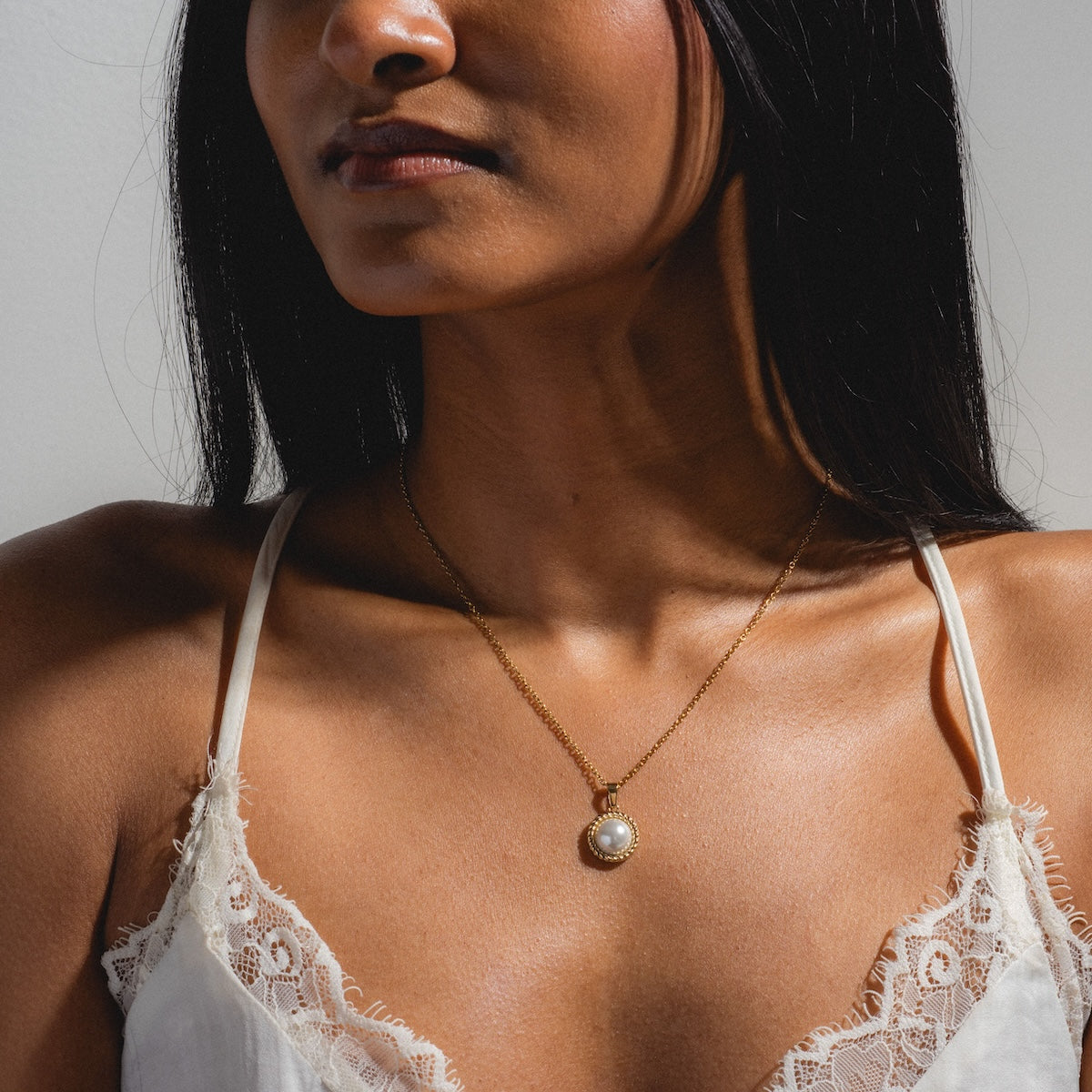 A woman wears a white lace-trimmed camisole and the Paloma Necklace, featuring a yellow gold-plated round pendant. The image focuses on her neck, shoulders, and upper chest in soft natural light.