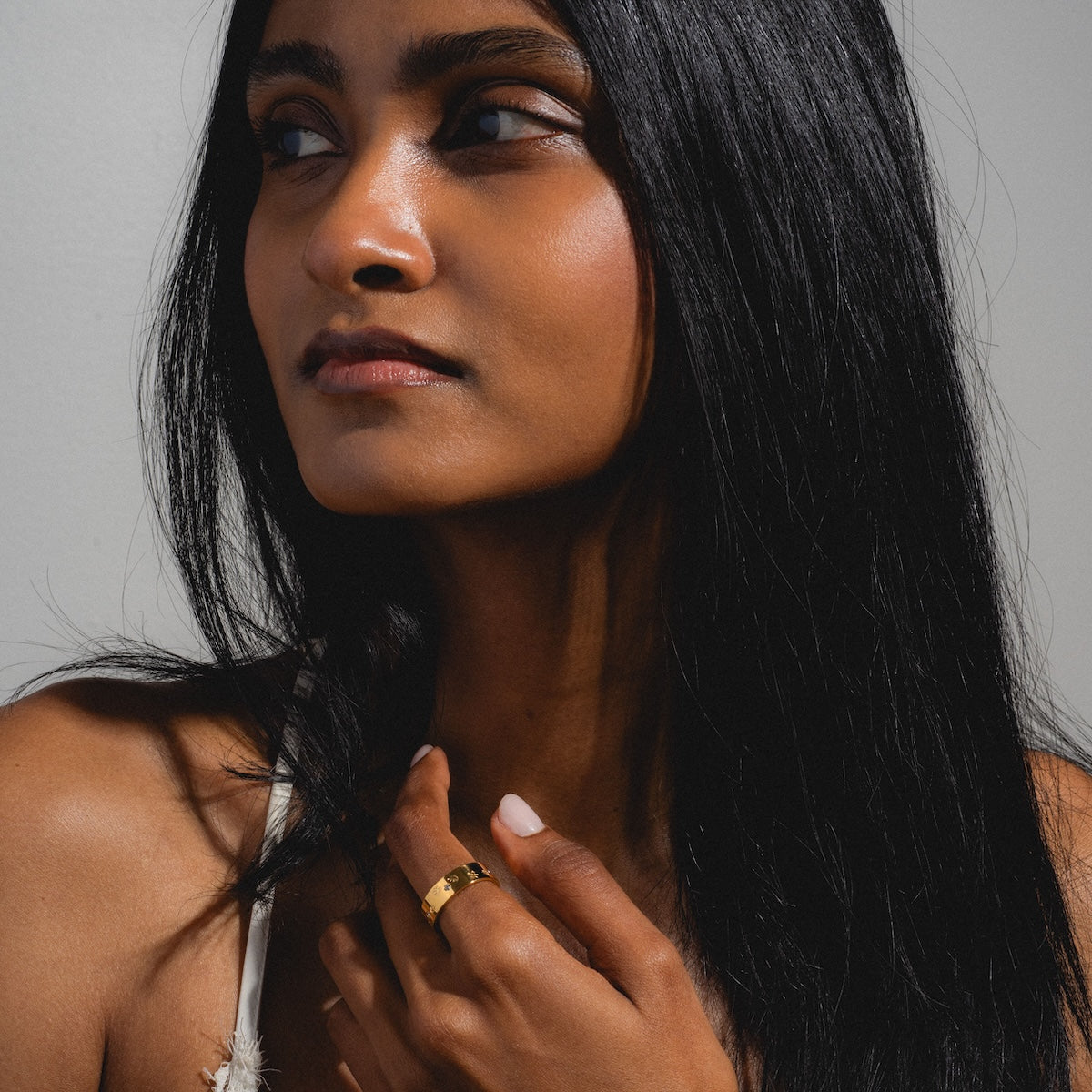 A woman with long black hair and a neutral expression gently touches her collarbone, wearing the Work of Art Ring in yellow gold plating with CZ stones and a white strap, standing against a plain gray background.