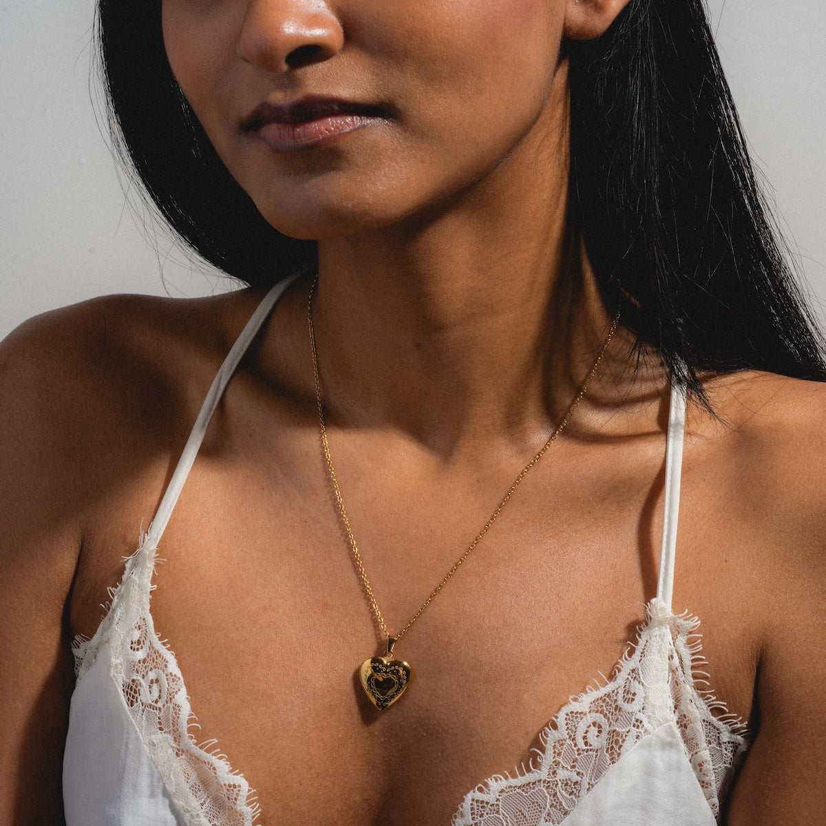 A woman wears a white lace camisole and the Daydream Necklace, featuring a gold heart-shaped pendant, her long dark hair resting on her shoulders against a simple background.