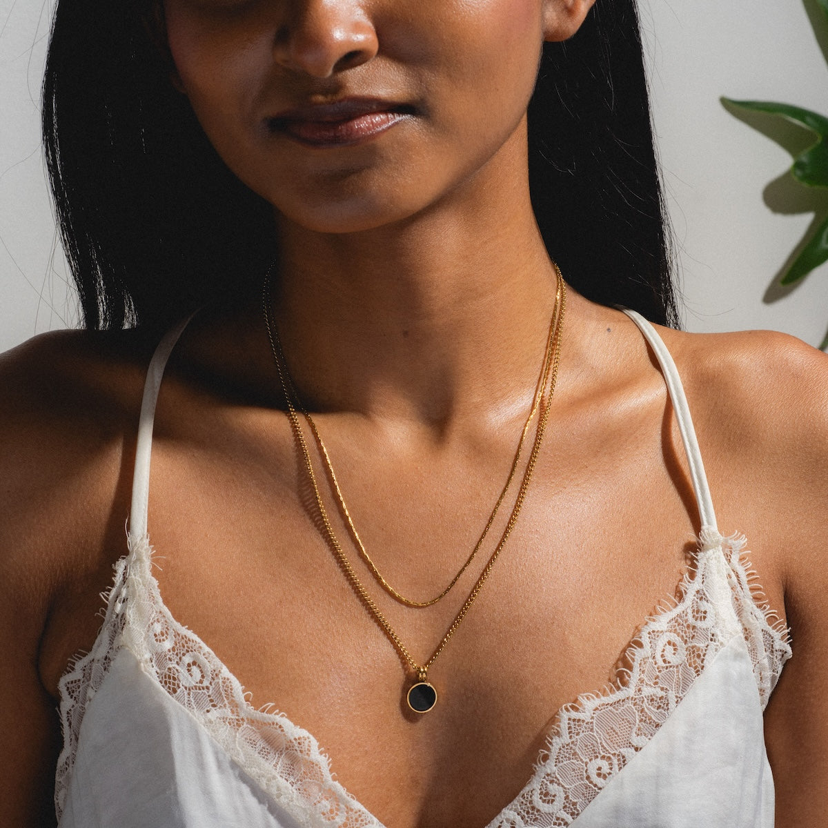 A woman wears a white lace camisole and the Billie Necklace with a black coin pendant, shown from the shoulders up. Sunlight creates shadows, and a green plant appears in the background.