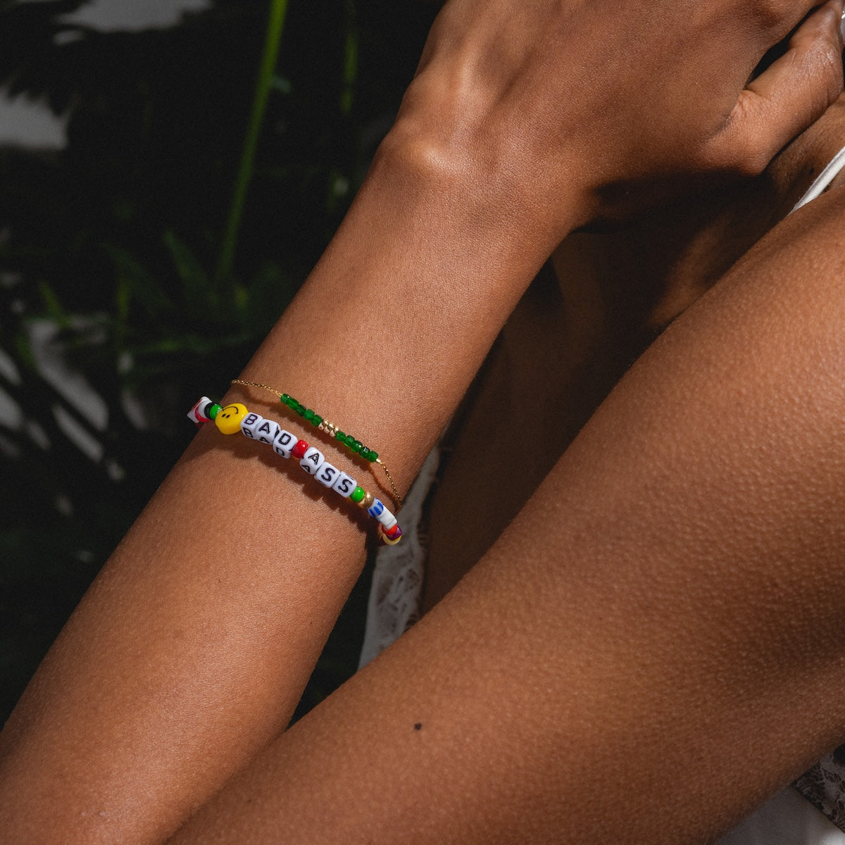 A close-up of a forearm wearing the Handmade Beaded Bada$$ Bracelet, featuring colorful beads and letter charms. The person wears a white sleeveless top with greenery blurred in the background.