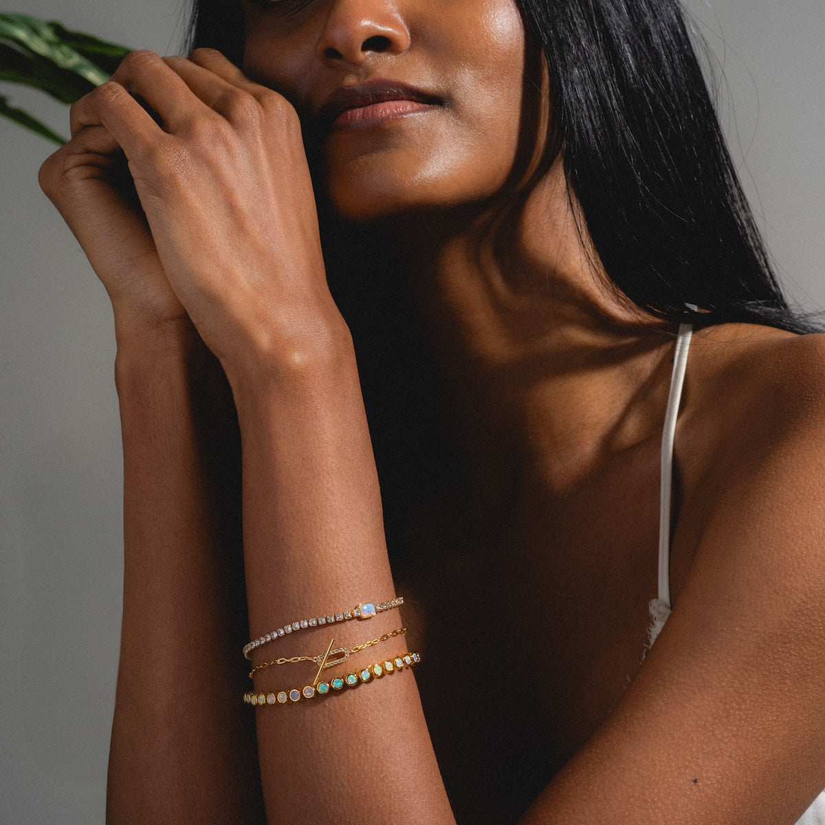 A woman with long dark hair wears a white camisole and the Tennis Bracelet with Opal, resting her chin on her hands and gazing softly away in natural light.