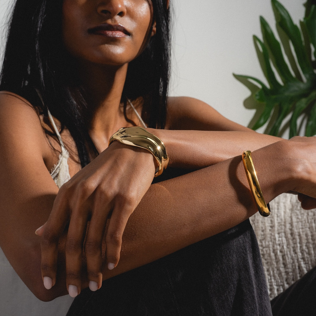 A woman with long dark hair sits with her arms crossed, wearing the Enza Small Bangle. She is dressed casually in a tank top and dark pants, with a leafy green plant in the background.