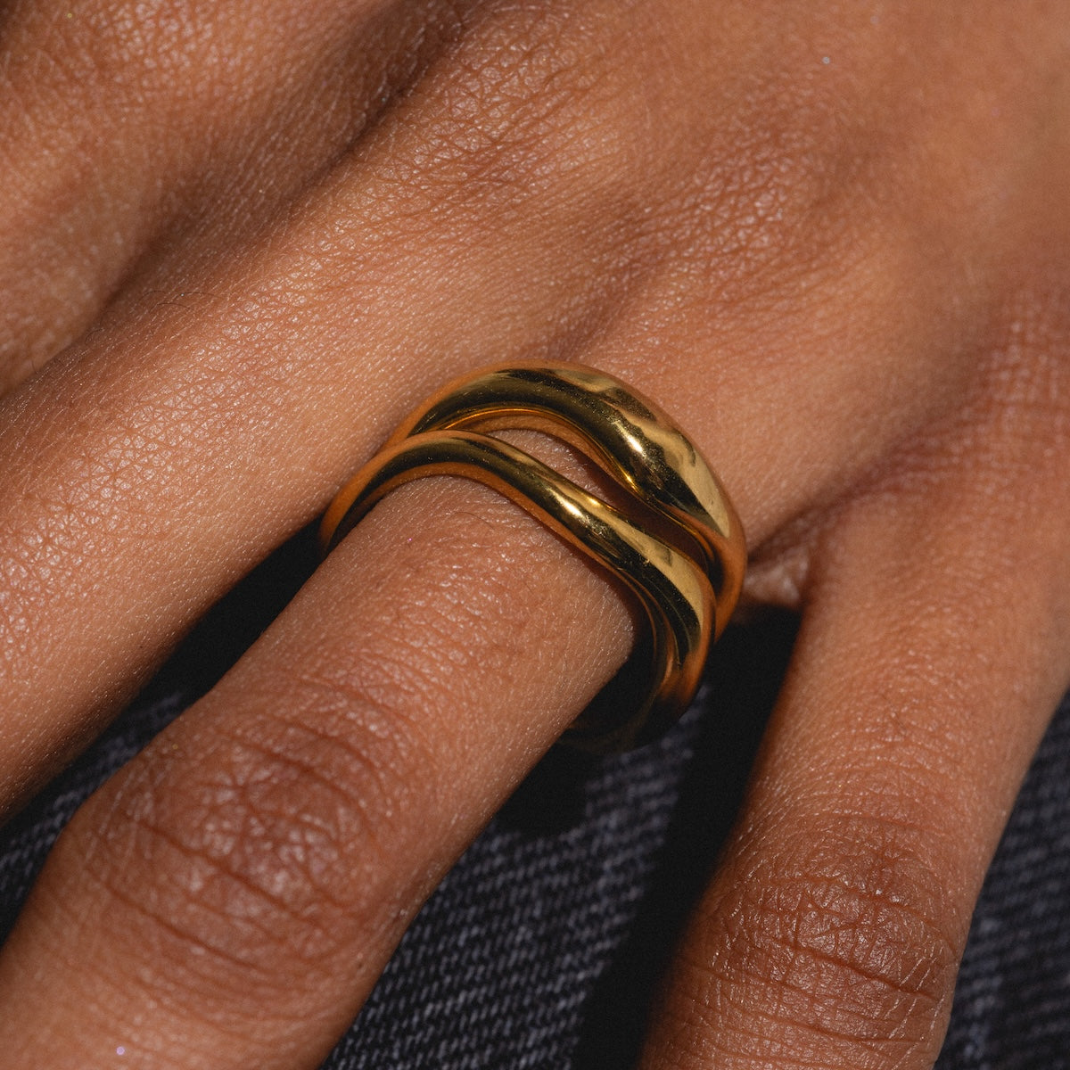 A close-up of a hand with a dark skin tone wearing the gold plated Curves Ring Set on the middle finger, set against a textured fabric background.