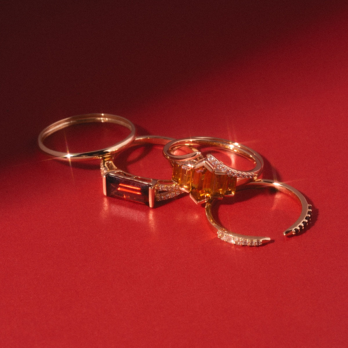 14k Delicate Yellow Gold Wedding Band, White Sapphire Open Front Nesting Ring, Black and White Open Front Nesting Band, Gilded Garnet East West Baguette Ring, and Graduated Citrine Deco Ring laying on bold red background