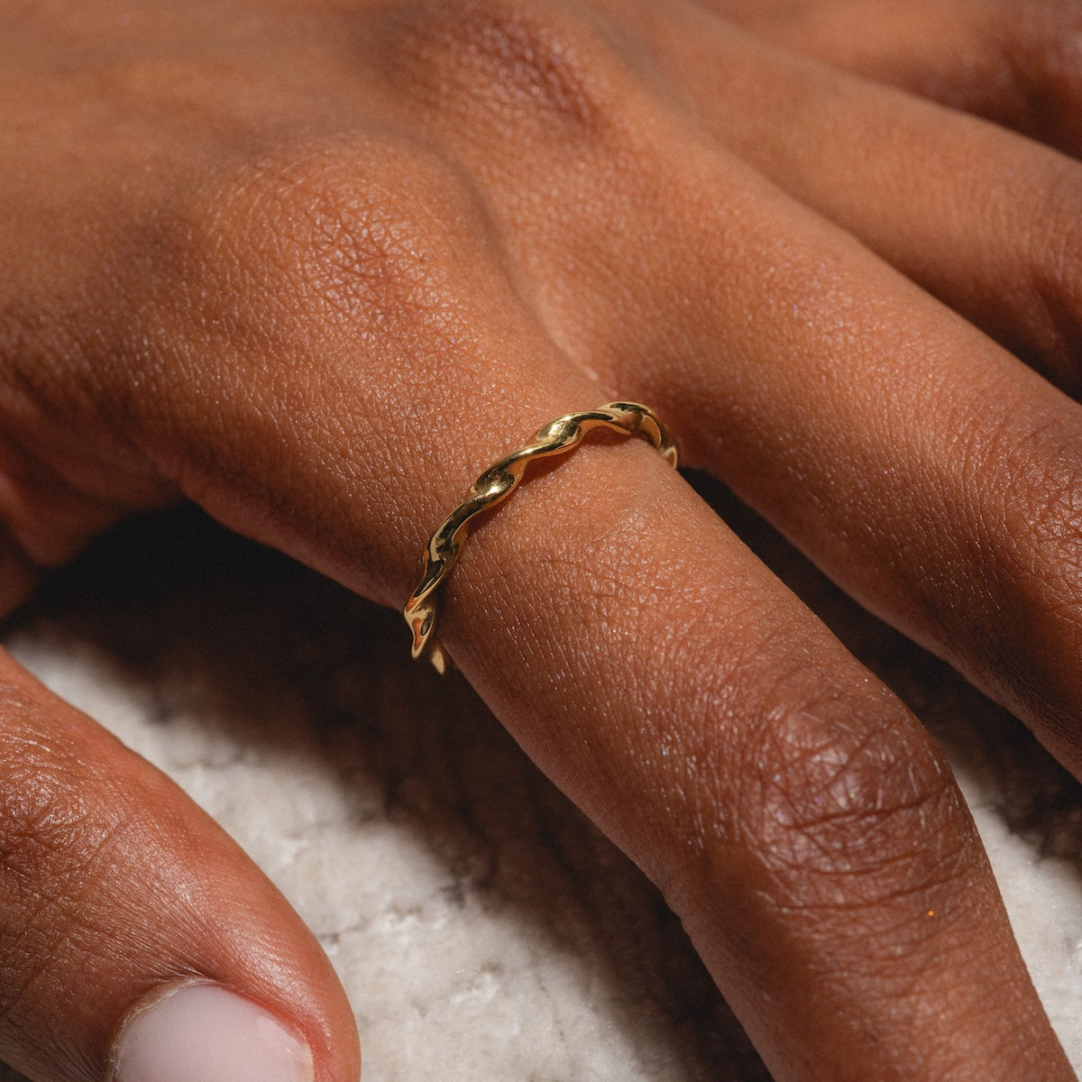 A close-up of a hand wearing the Souvenirs Ring, a delicate yellow gold piece with a twisted band, on the ring finger. The hand rests on a light textured surface as natural lighting highlights the ring’s shine.