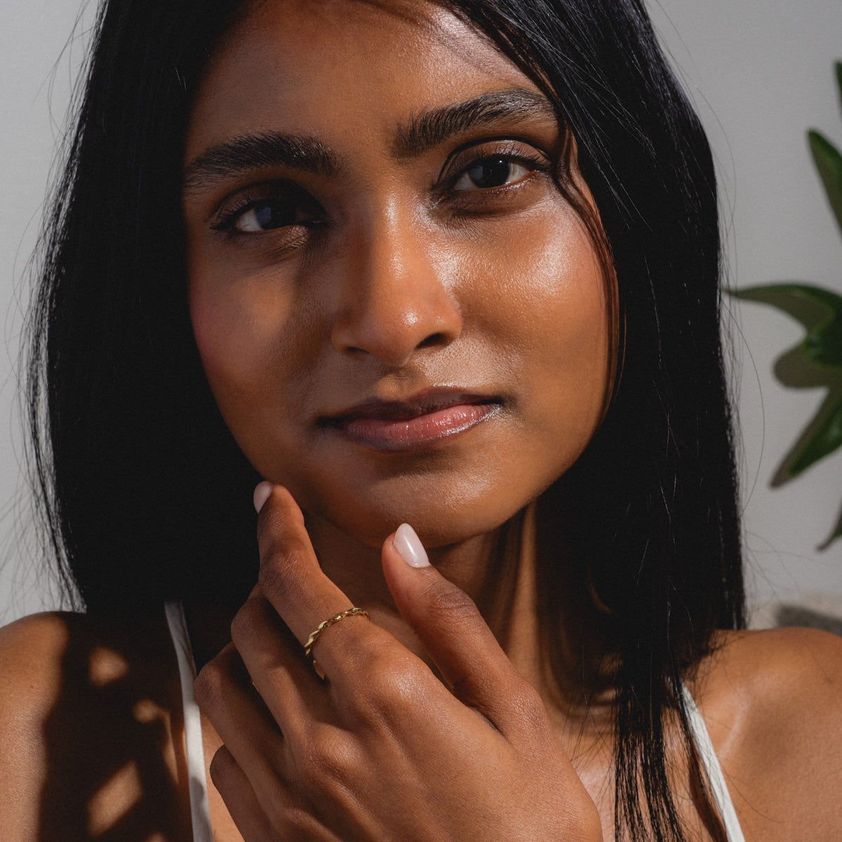 A woman with long dark hair gazes at the camera, touching her chin and showcasing the Souvenirs Ring in yellow gold. She has smooth skin, natural makeup, sunlight plays across her face, and a green plant appears in the background.