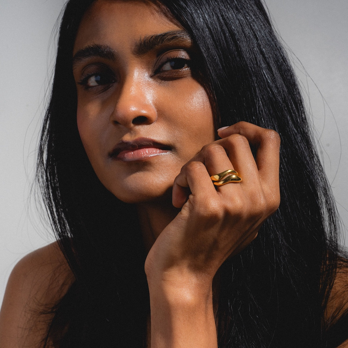 A woman with long dark hair gazes thoughtfully to the side, her chin resting on her hand adorned with the elegant Curves Ring Set. She wears a serene expression against a neutral background.