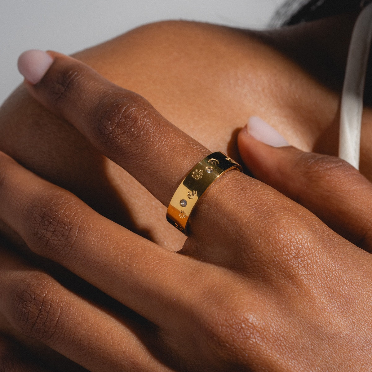 A close-up of a hand with medium brown skin wearing the Work of Art Ring, a yellow gold plated engraved band with small symbols, resting gently on a bare shoulder. The nails are manicured in soft pink polish.