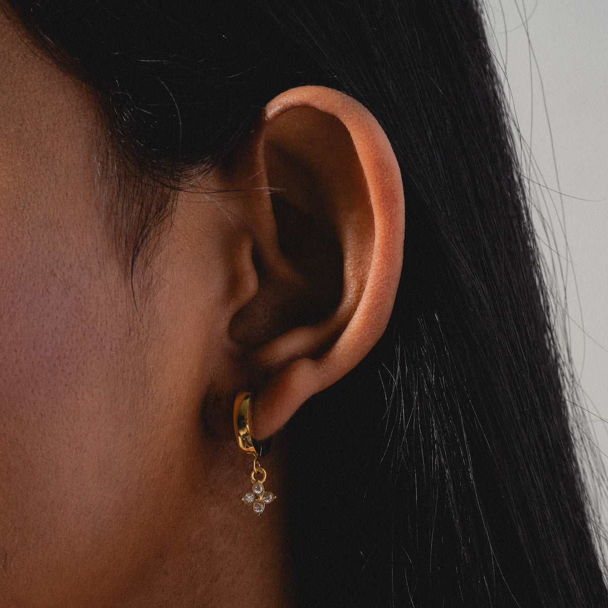 A close-up of a person with long dark hair wearing Merveilles Hoops—gold huggie earrings featuring a dangling flower-shaped charm embellished with tiny crystals.