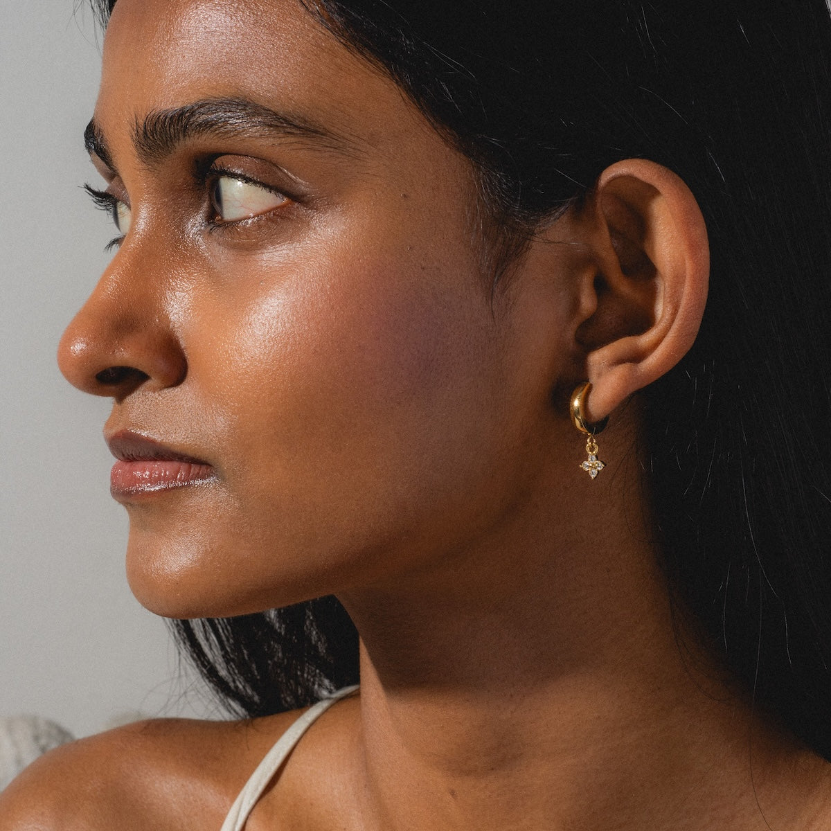 A woman with smooth brown skin and straight black hair looks to the side, wearing gold Merveilles Hoops with a small star charm. A cream-colored strap is visible on her shoulder against a plain, light background.