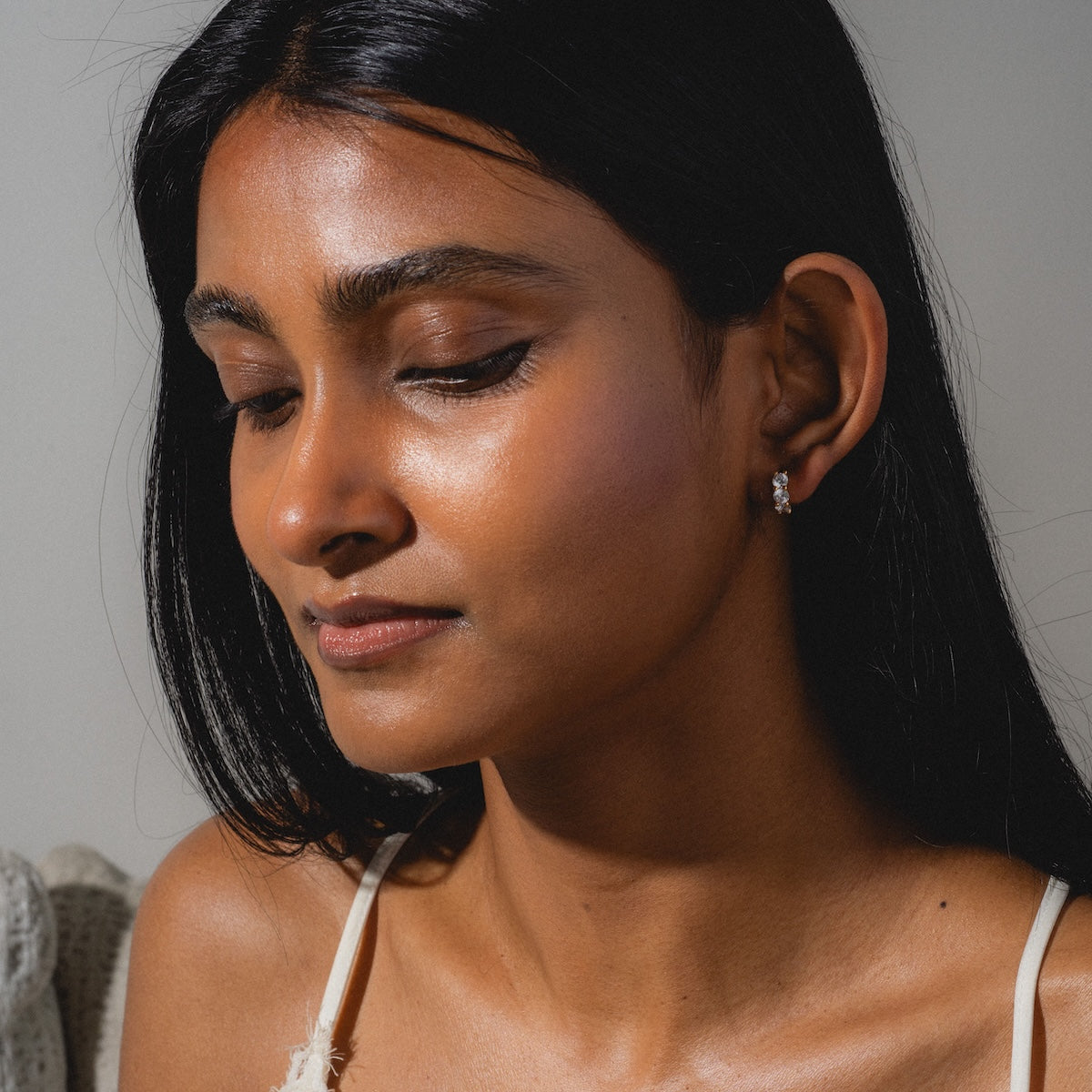 A woman with long dark hair and glowing skin looks down calmly, wearing a white top and Eclat Hoops. Soft light highlights her face against a neutral background.