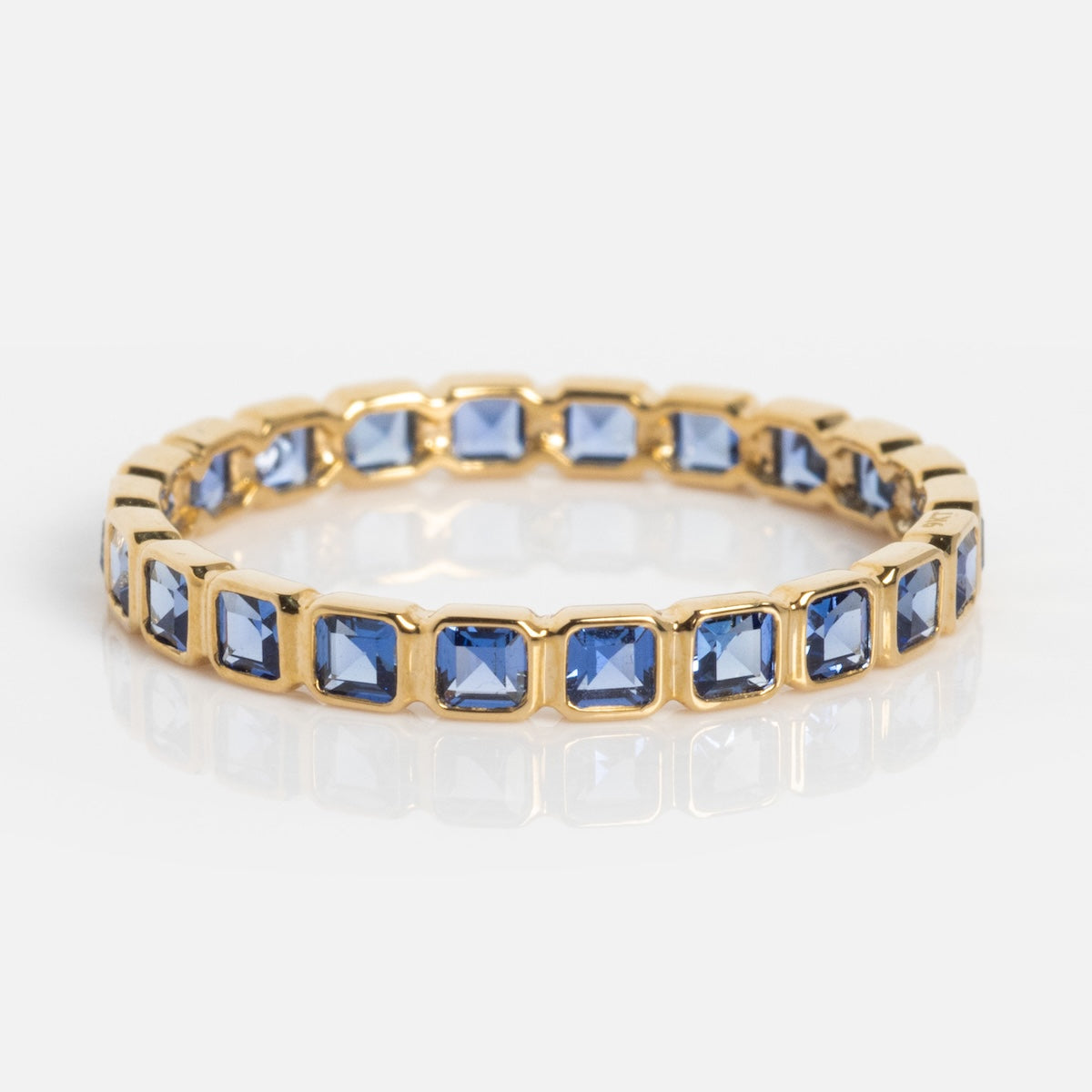 The Solid Gold Gemstone Eternity Stacking Ring features square-cut blue gemstones in prong settings, ideal for stacking, and is shown on a reflective white surface.