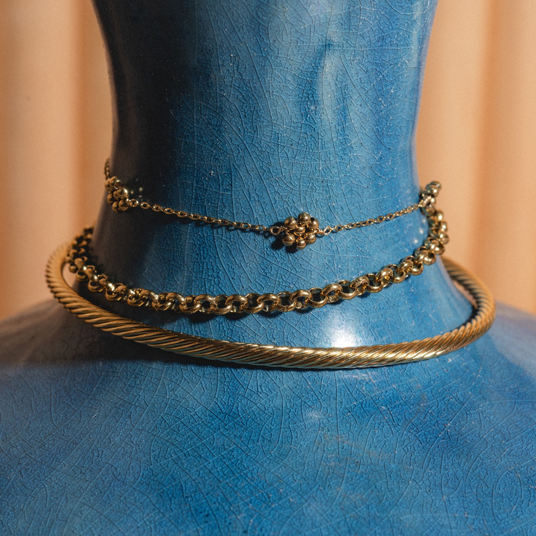 A blue mannequin bust displays the Ampthill Bangle alongside two gold necklaces—a delicate 14k gold filled chain with a small cluster pendant and a medium round link chain—with beige fabric in the background.