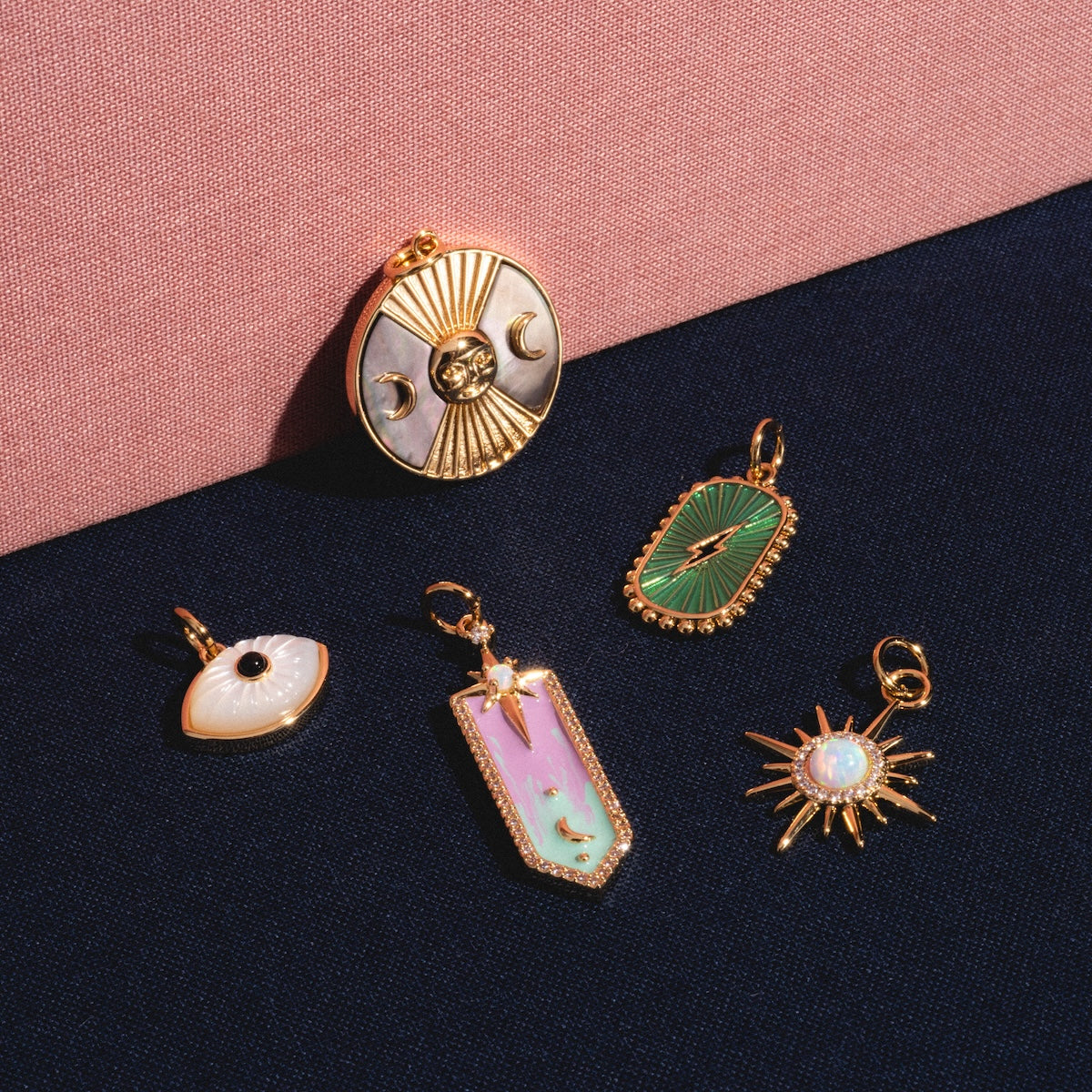 Five Charms by Elizabeth Stone, featuring celestial and eye motifs in 14k gold plated metal with colorful enamel and mother of pearl, are displayed on a dark blue surface with a pink textured background.