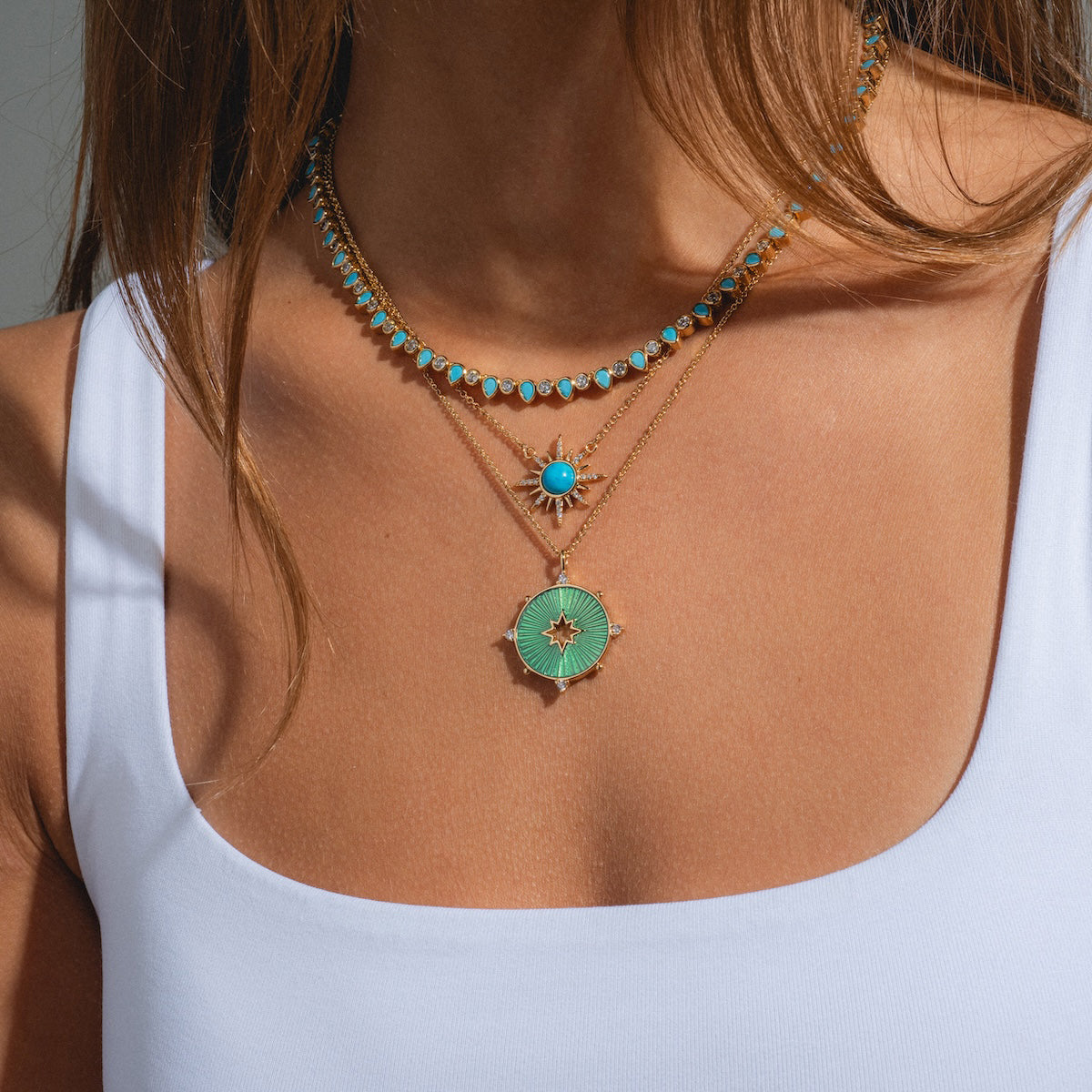 A woman wears a white tank top and three layered gold necklaces, including the Teardrop Tennis Necklace with cubic zirconia, plus two more with turquoise and green pendants—one detailed with small turquoise beads.