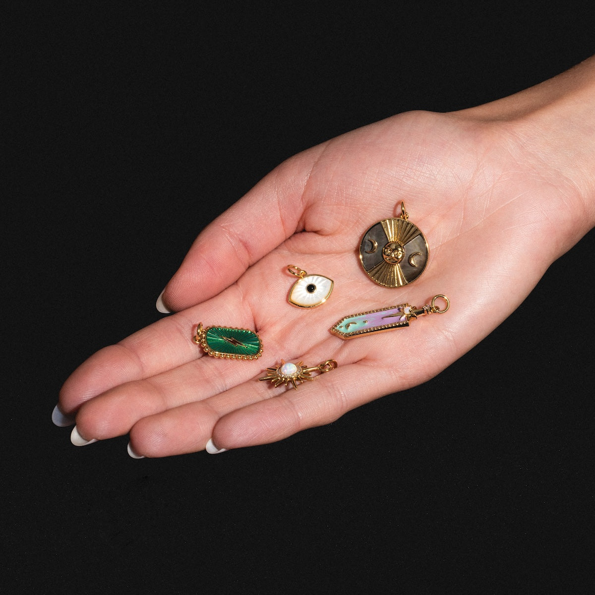 A manicured hand displays six Charms by Elizabeth Stone—14k gold plated celestial charms, green, white mother of pearl, purple stones, and a round gold charm—all set against a black background.