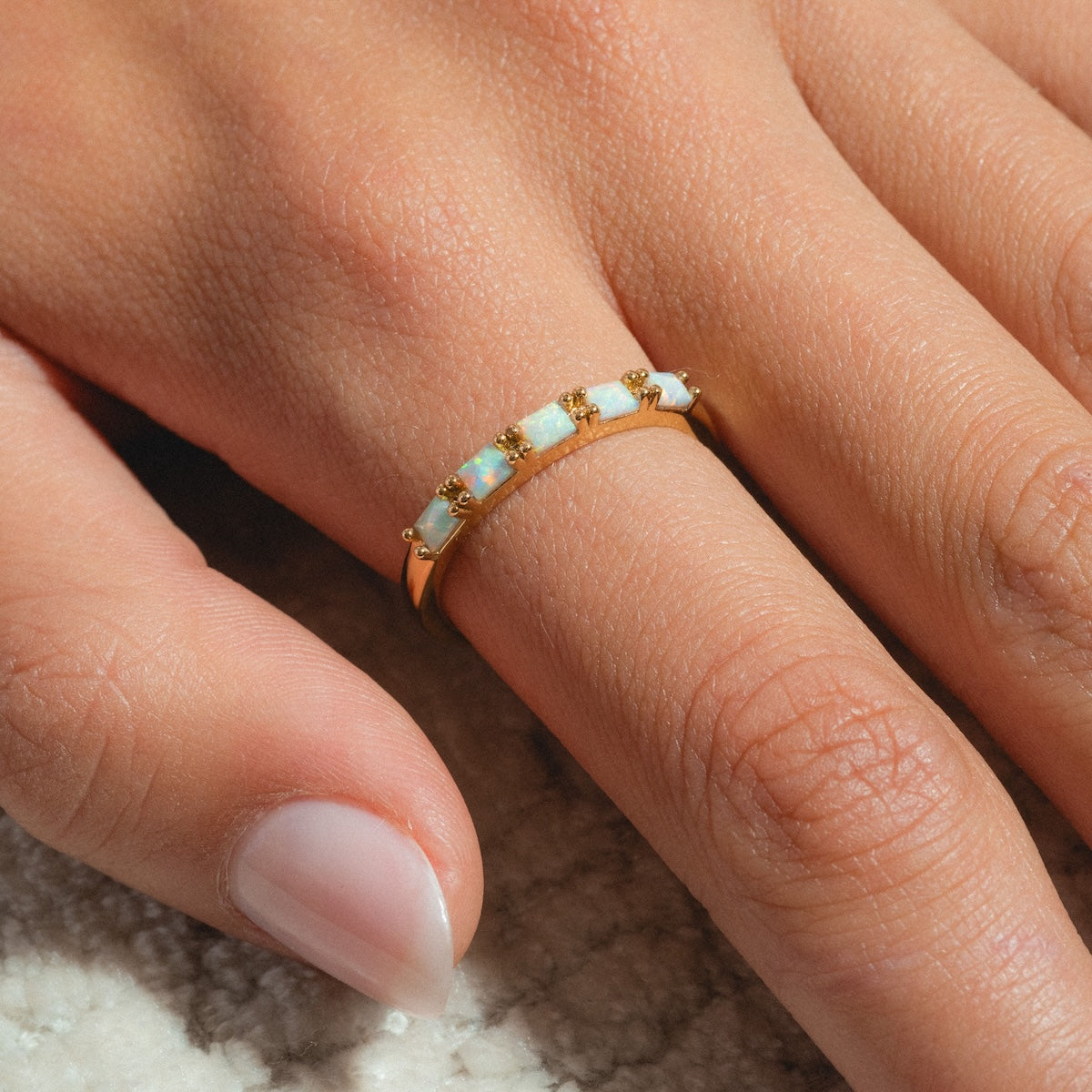 A close-up of a hand wearing the Opal Baguette Band, a 14k gold plated ring set with five opal stones, on a textured surface. The nails are natural and glossy—ideal for stacking or wearing solo.