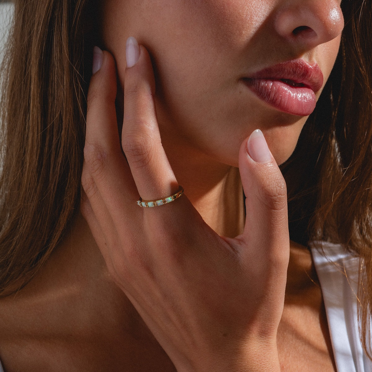 A close-up of a woman with natural makeup and manicured nails gently touching her cheek, showcasing the elegant Opal Baguette Band on her finger.