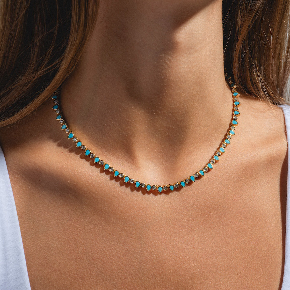 A woman wears the Teardrop Tennis Necklace, a delicate 14k gold piece with small turquoise stones, shown close-up against her bare neck and collarbone with a white top at the edges.