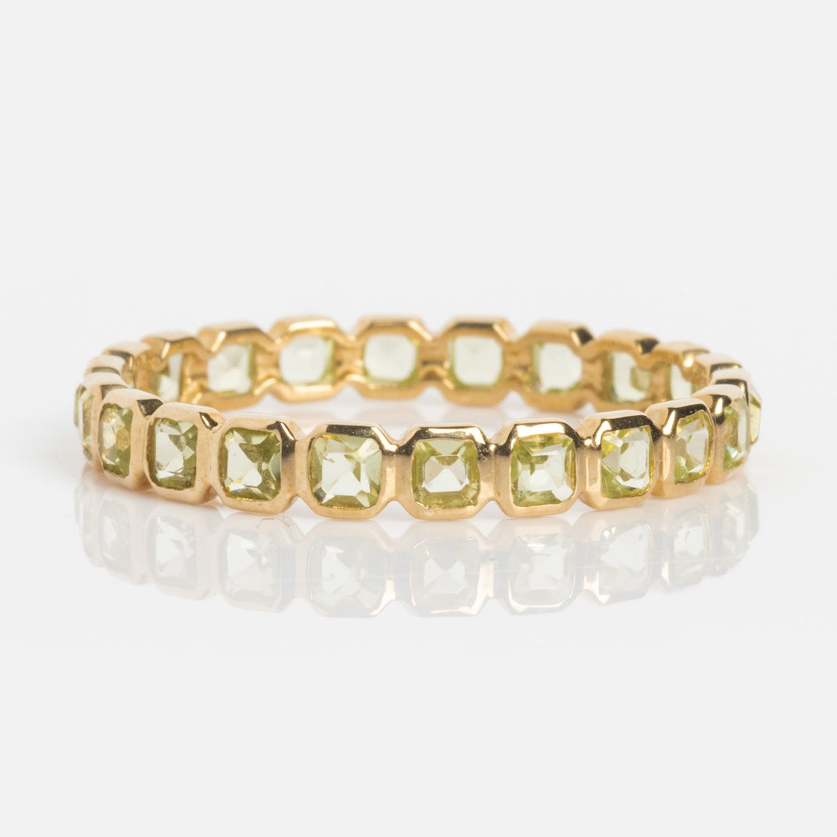The Solid Gold Gemstone Eternity Stacking Ring features square pale green peridot stones and is shown on a white reflective surface.