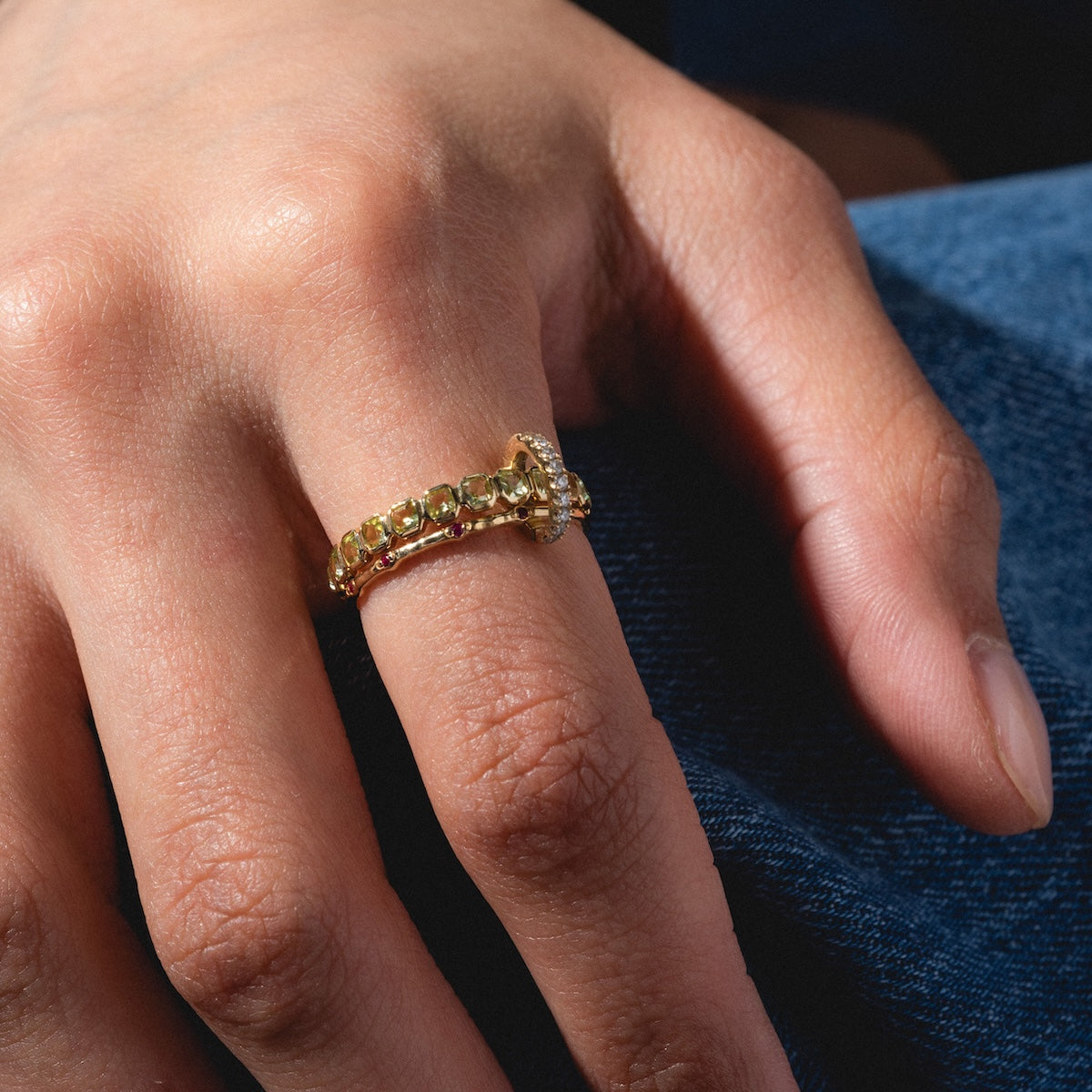 A close-up of a hand on denim wearing the Solid Gold Ring Clip stack—two gold rings: one with small green gemstones, the other with a larger clear gemstone—showing customizable style on the ring finger.