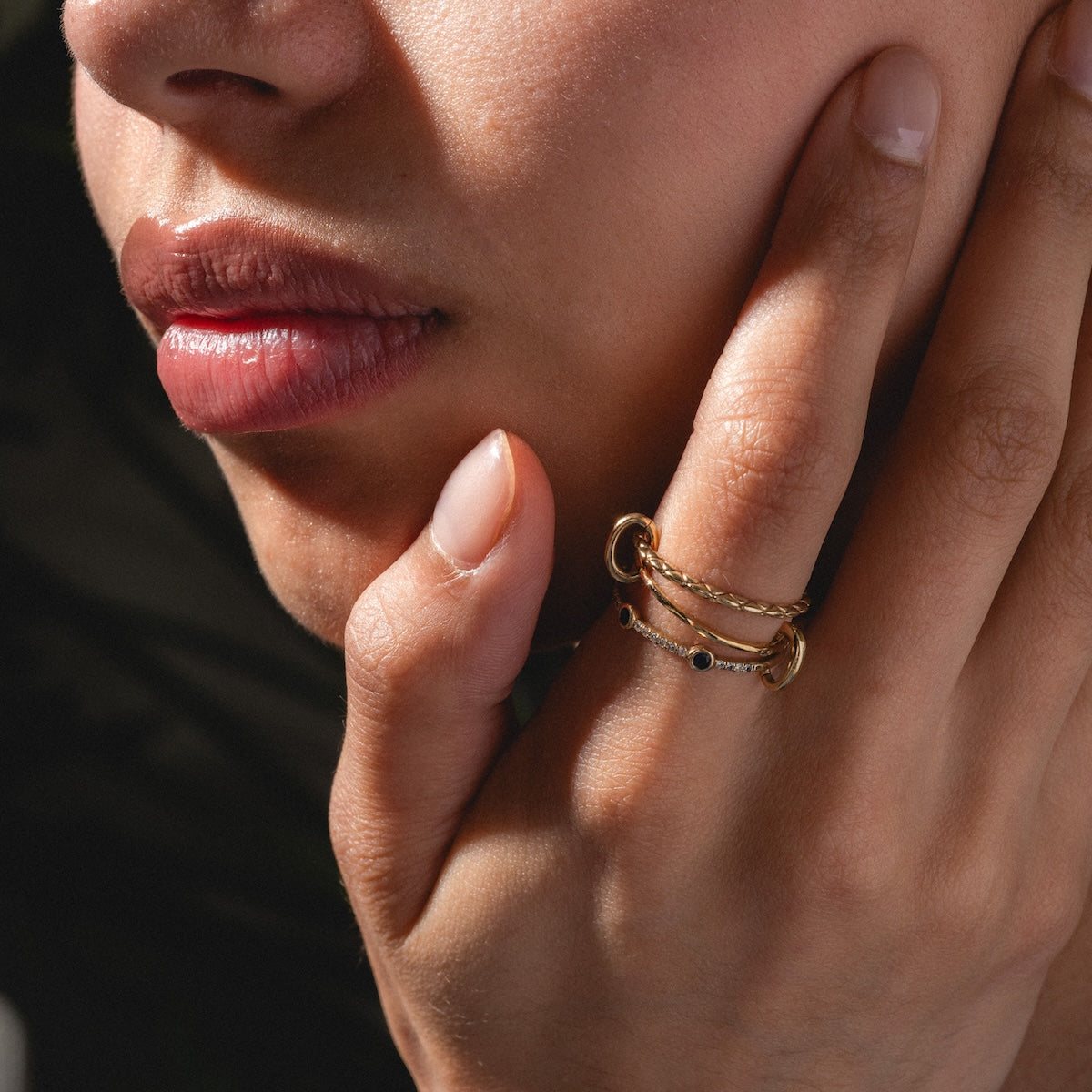 Close-up of a person touching their face, highlighting the Solid Gold Ring Clip stacked with other gold rings and black stones. The lighting emphasizes the skin texture and fine jewelry details.