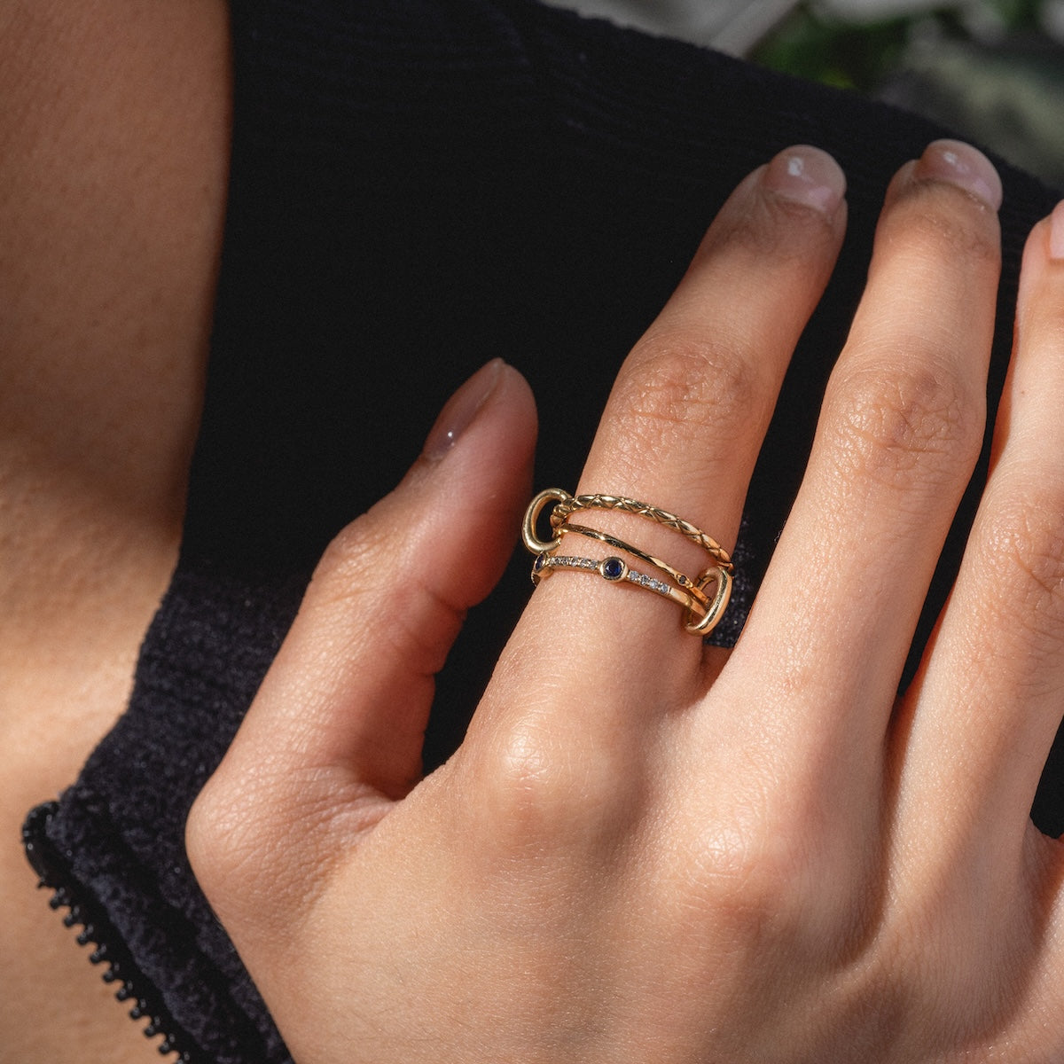 A close-up of a hand wearing the Solid Gold Ring Clip—two connected gold bands with a small dark stone—styled in a customizable ring stack. The person has natural nails and wears a dark zip-up garment.