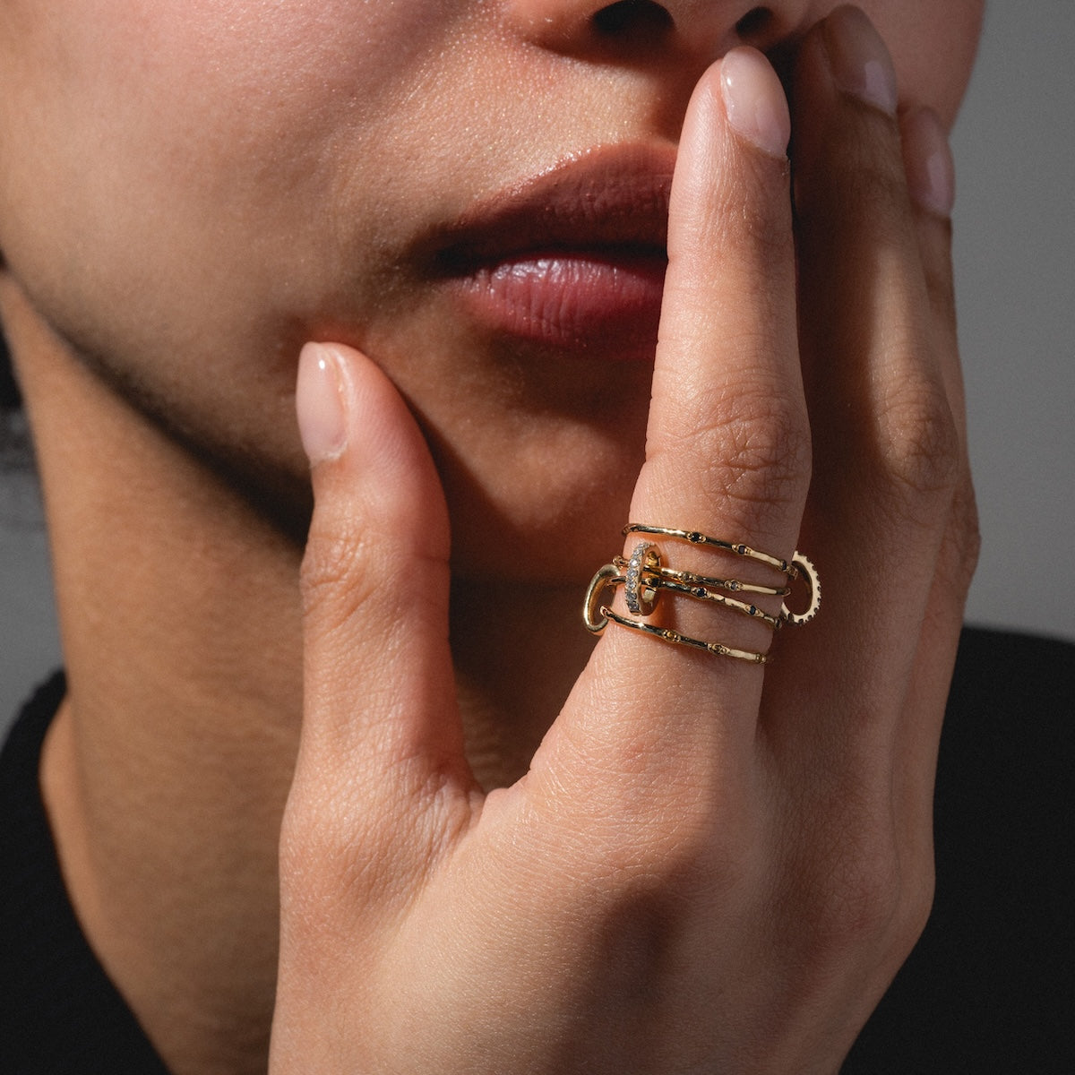 Close-up of a person touching their lips, showcasing the Solid Gold Ring Clip styled in a unique DIY ring stack. Multiple gold bands adorn their fingers against a neutral background, and theyre wearing a dark top.