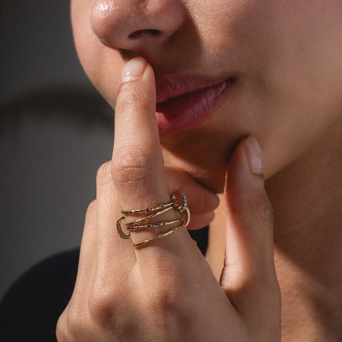 A person with soft pink lips gently touches their face, highlighting the Solid Gold Ring Clip—a large, intricate, customizable stackable ring with multiple bands and textured details—against a blurred background.