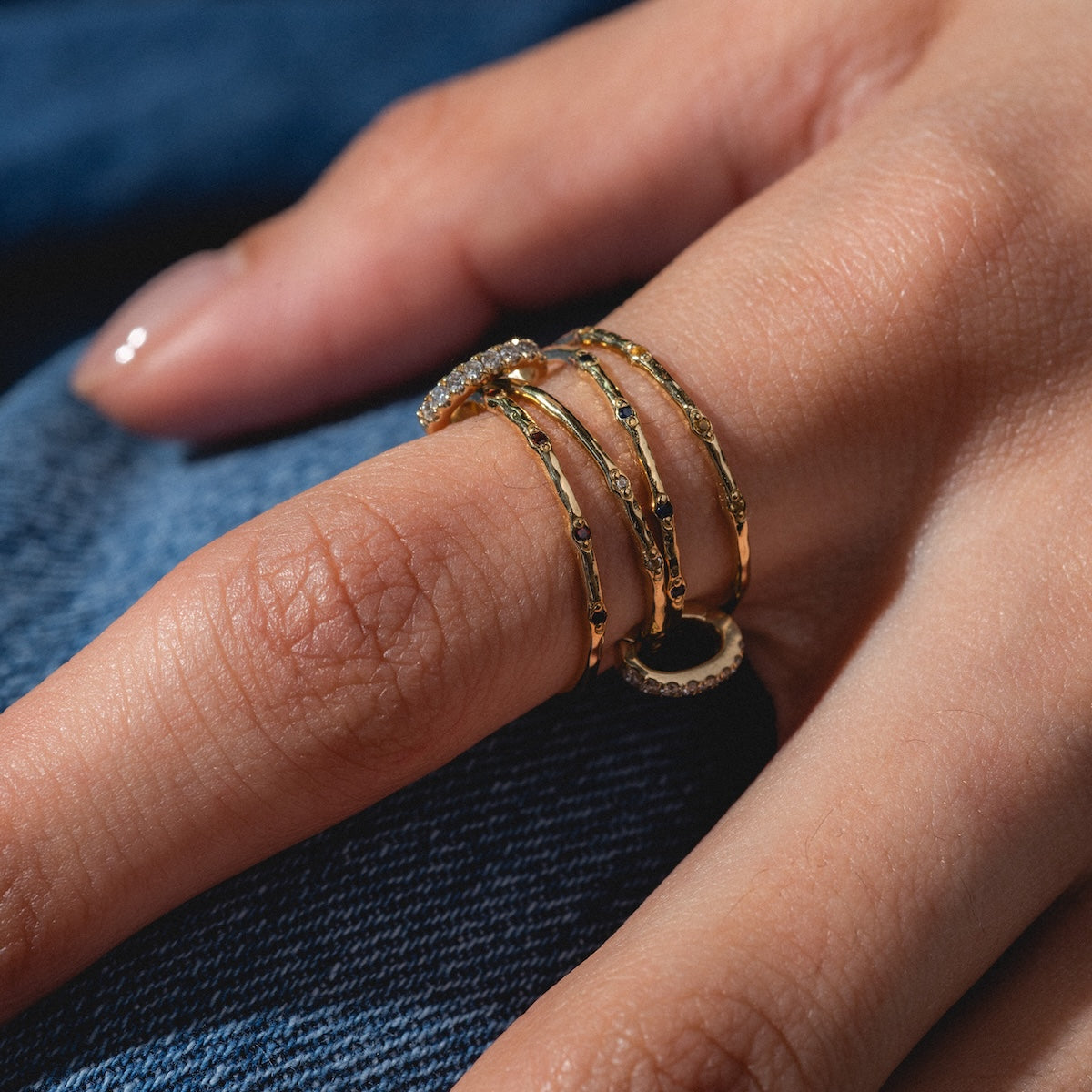 A close-up shows a hand wearing four Solid Gold Ring Clips—some with small dark stones, one with clear stones—stacked on the middle finger, set against blue denim fabric.