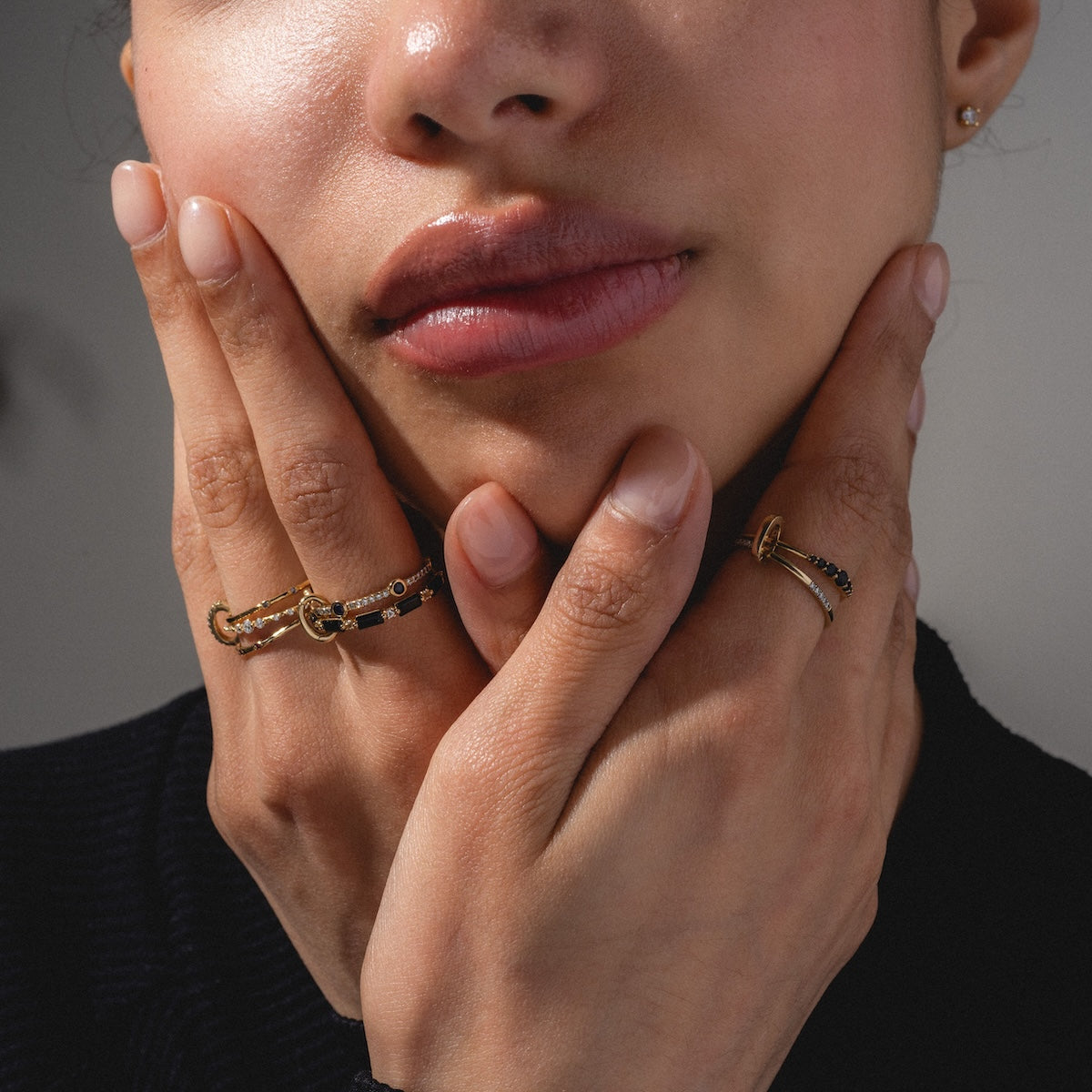 A close-up of a womans lower face and hands showcases the Solid Gold Ring Clip styled in a DIY ring stack with black stones; her fingers gently touch her cheeks, emphasizing natural makeup and a stud earring.