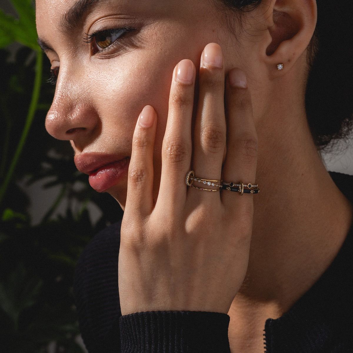 A woman with smooth skin rests her hand on her cheek, displaying a DIY stack of three Solid Gold Ring Clips. She wears a dark top and small stud earrings, with green leaves and soft lighting in the background.