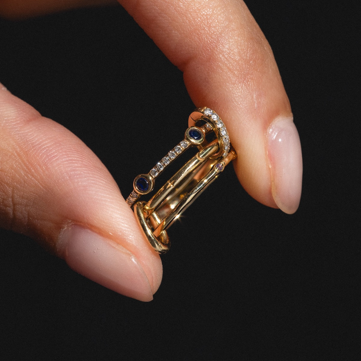A manicured hand holds the Solid Gold Ring Clip, adorned with two blue gemstones and a row of clear crystals—perfect for creating your own customizable ring stack—set against a black background.