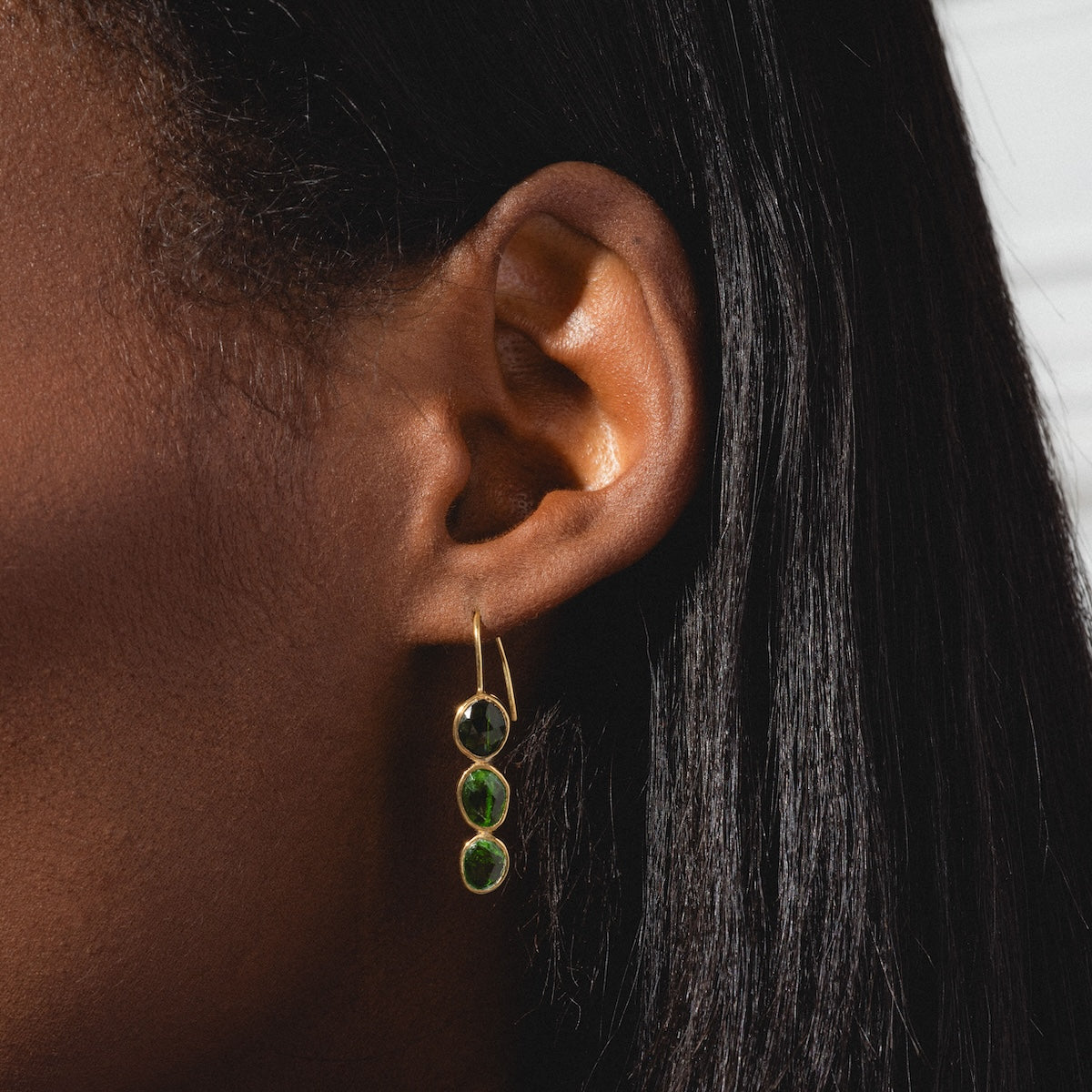 A close-up shows a person with dark skin and straight black hair wearing Amber Earrings—yellow gold plated with three round green tourmalines, each bezel set and hanging vertically.