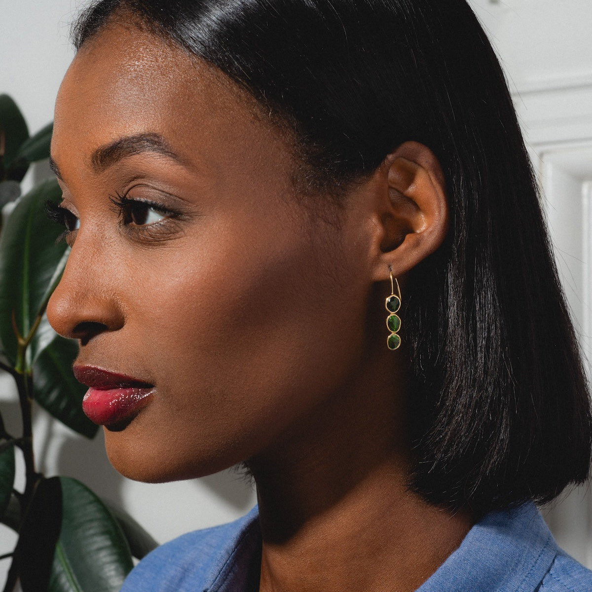 A woman with straight shoulder-length hair and red lipstick wears Amber Earrings, yellow gold plated with green tourmaline stones, shown in profile against a white wall and green-leafed plant.