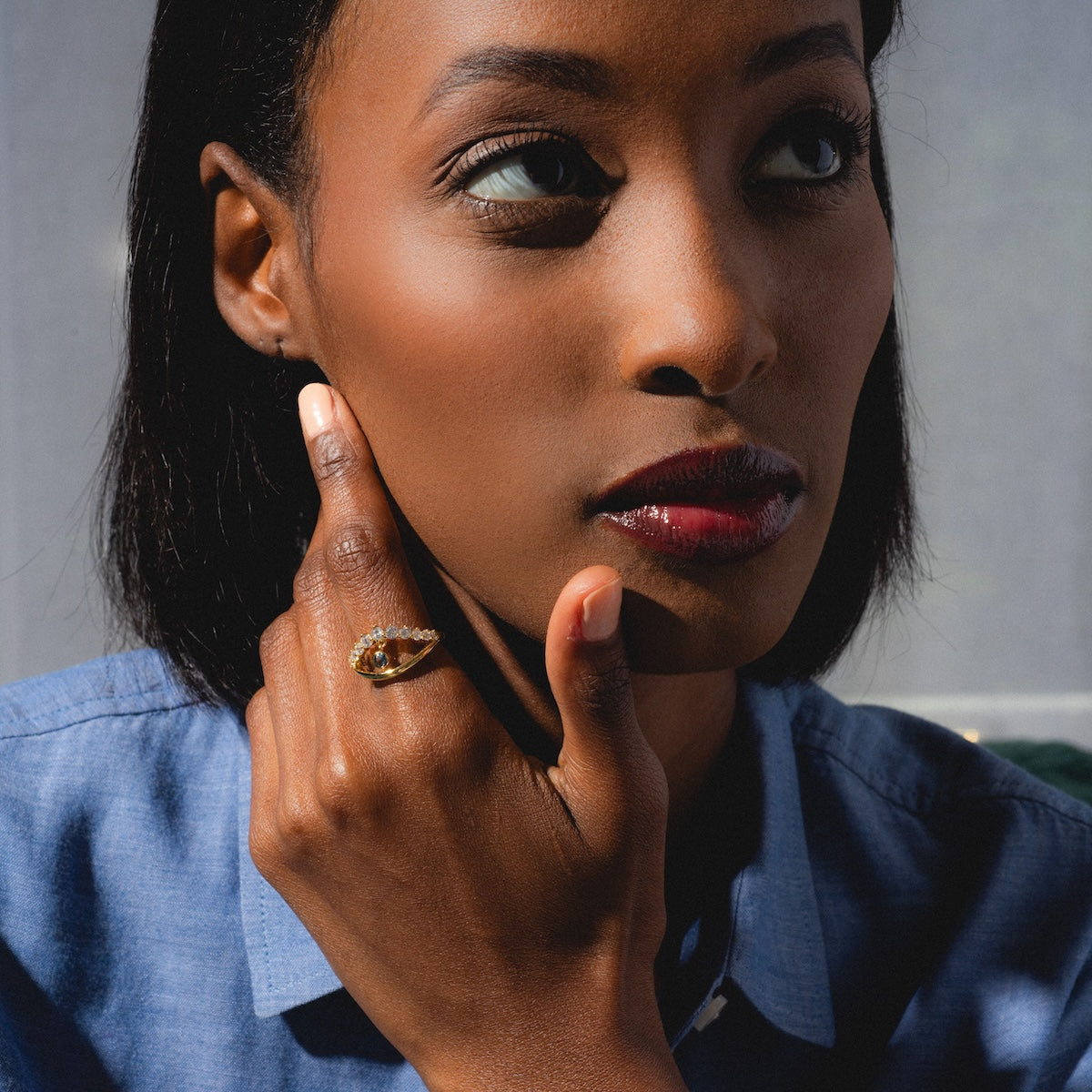 A woman in a blue shirt gazes thoughtfully to the side, her chin resting on her hand as she wears the Mystique Evil Eye Charm Ring, its 14k yellow gold plating and brown stone catching the sunlight against a muted background.