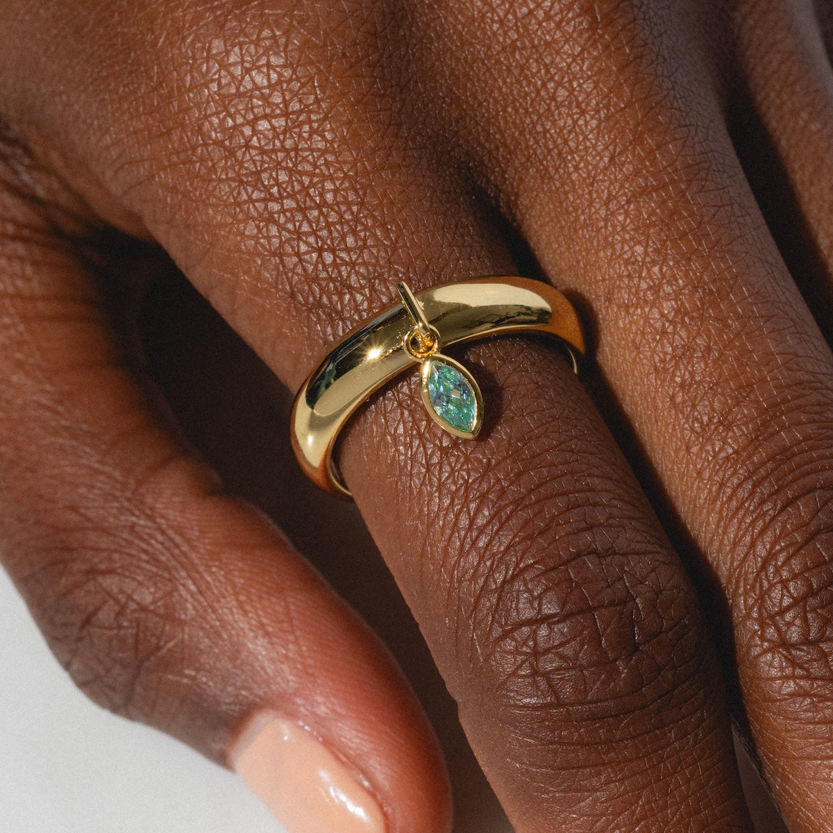 A close-up shows a hand with natural nails wearing two gold rings, including the Classic Shimmer Charm Ring with a teardrop green gemstone on the middle finger, highlighted by soft lighting.
