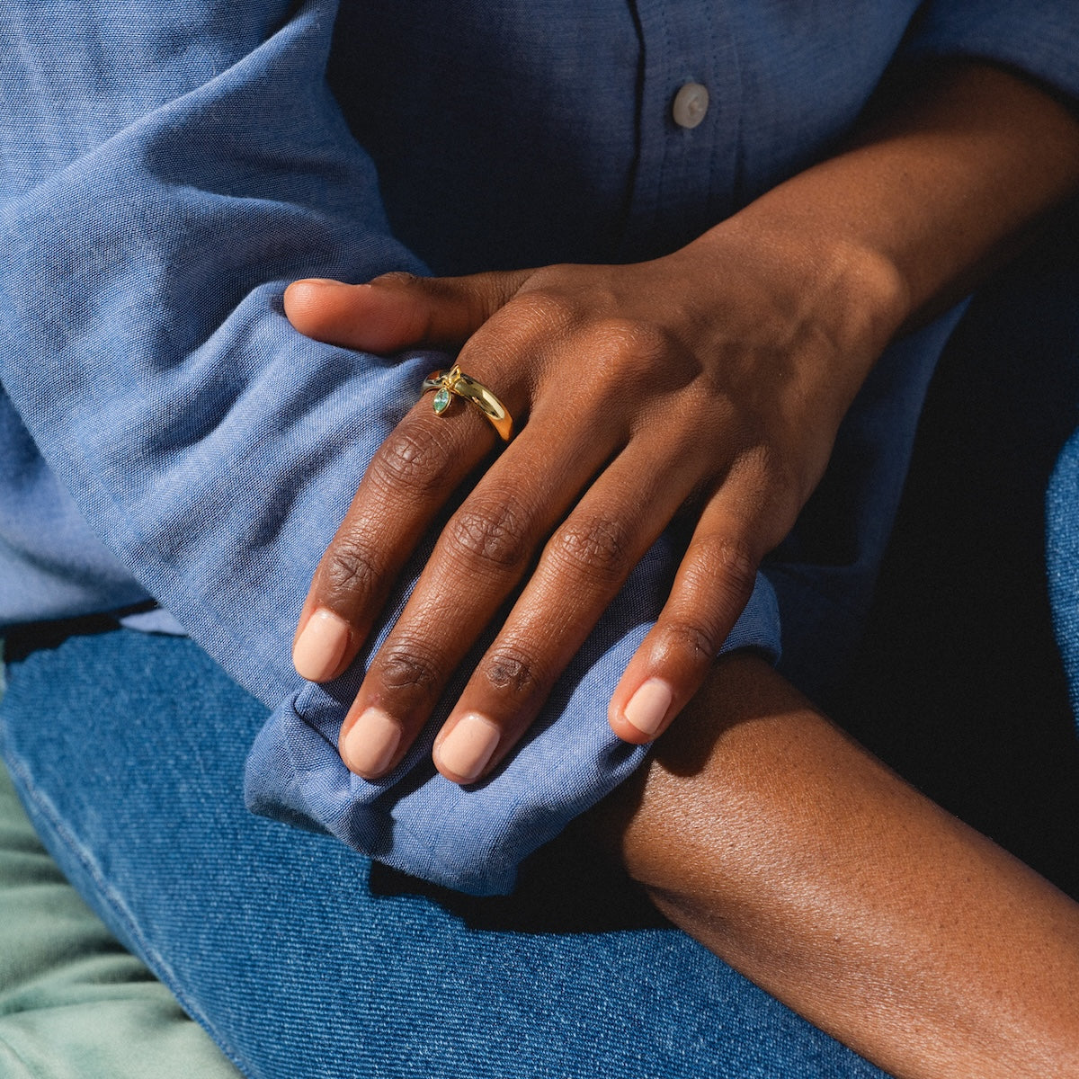 A person in a blue shirt and jeans rests their hand on their knee, displaying the Classic Shimmer Charm Ring—an exclusive gold band with a gemstone—on their finger and featuring neatly manicured, light pink nails.