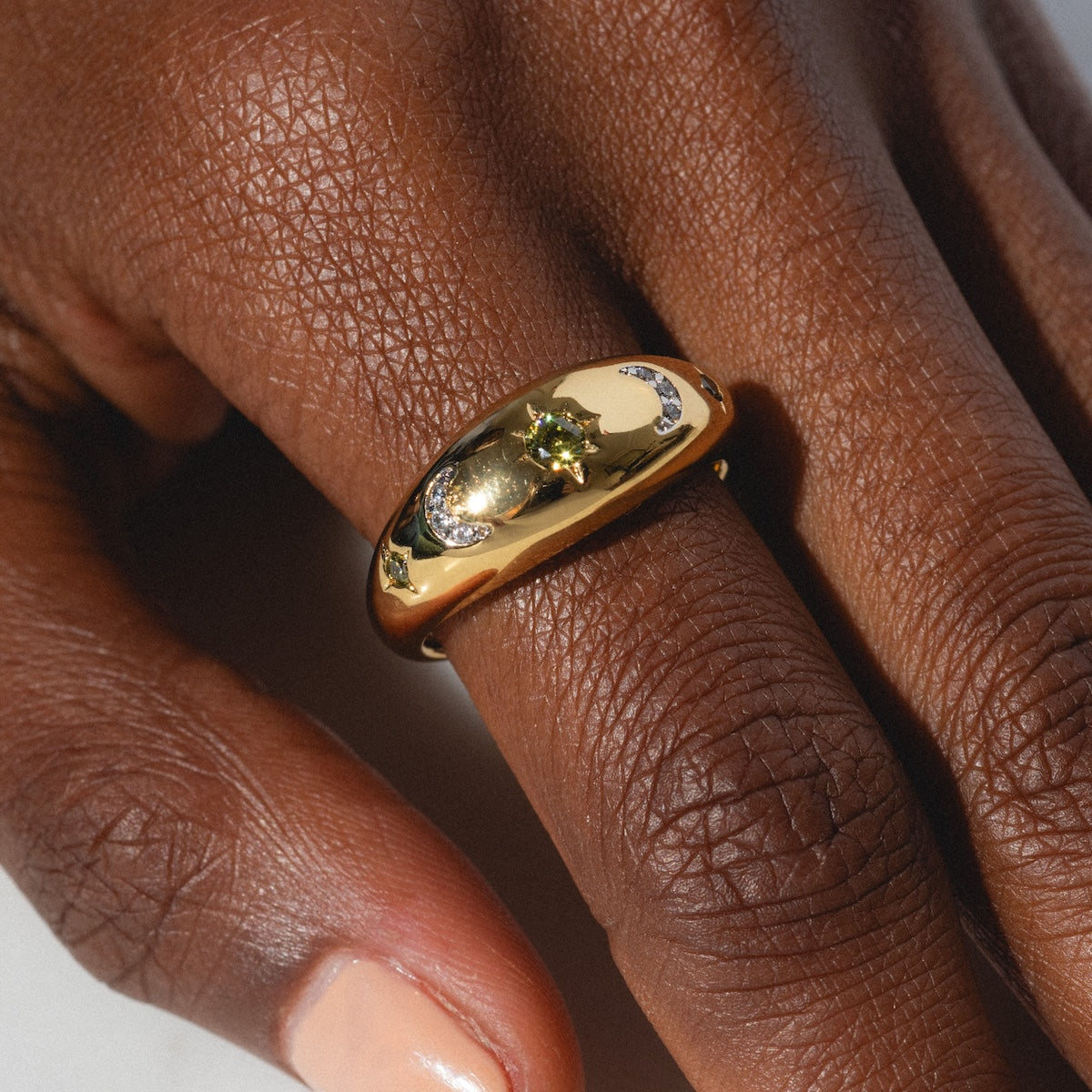 A close-up of a hand with medium-dark skin wearing the Moonstruck Bold Dome Ring, a 14k gold plated piece engraved with stars and moons and featuring a green gemstone at the center. Nails are neatly manicured with pale polish.