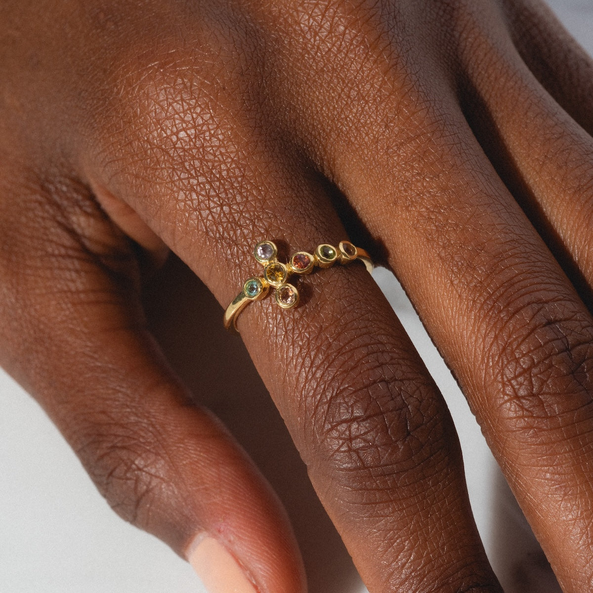 A close-up of a hand wearing the Notre Dame Ring, a yellow gold plated piece set with multiple small, colorful tourmaline gemstones, shown on the middle finger against a neutral light background.