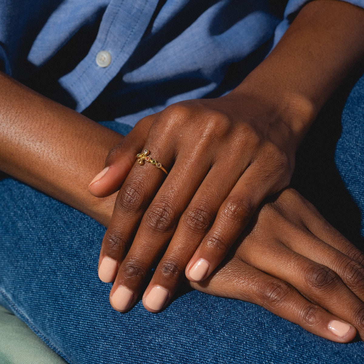 A person with neatly manicured, natural nails wears the Notre Dame Ring—a delicate yellow gold plated multi-color tourmaline ring—resting one hand over the other, dressed in blue jeans and a blue shirt.