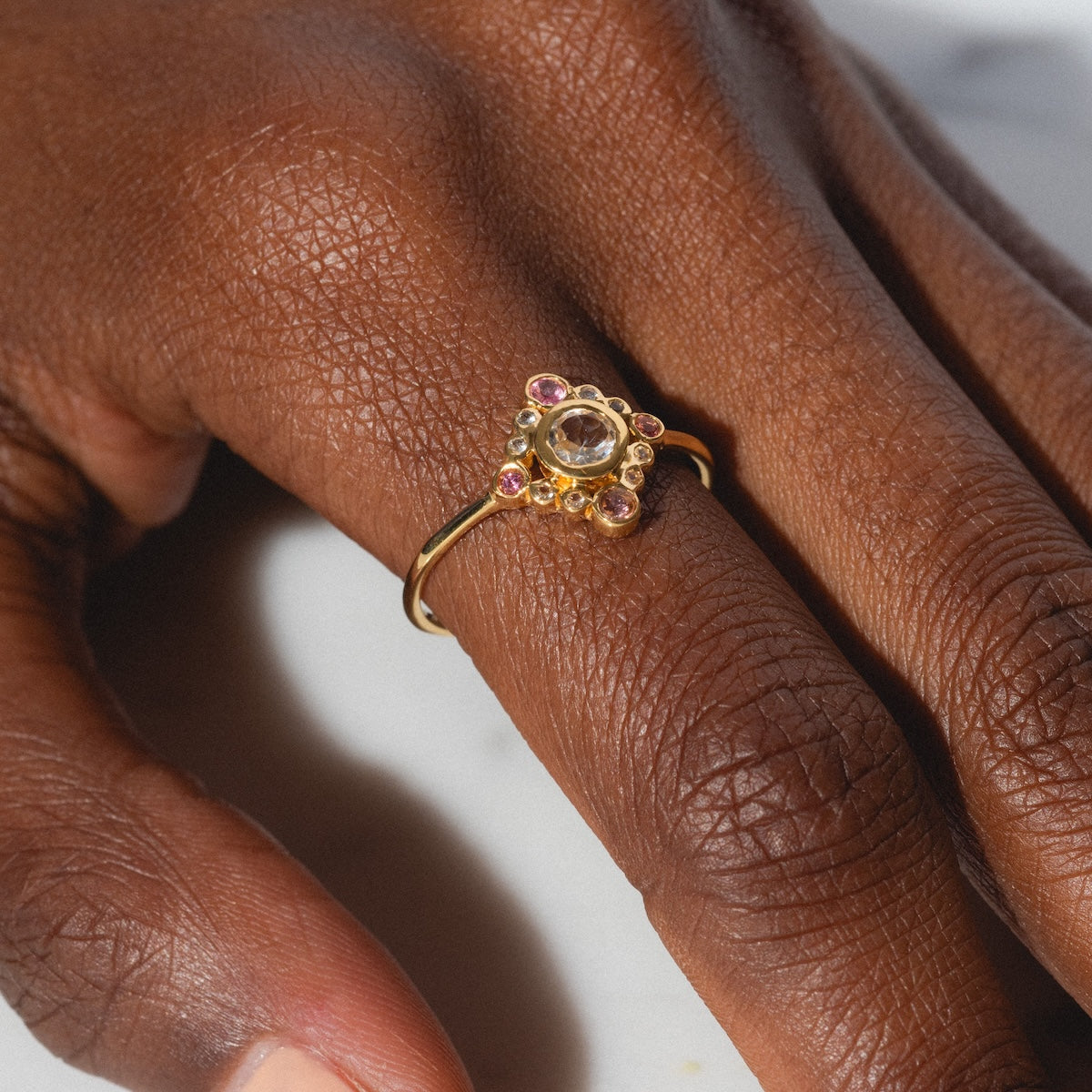 A close-up of a hand wearing the Oberoi Ring, a yellow gold plated piece featuring a central clear gemstone encircled by smaller pink and gold stones in a striking multi-stone design, set against a simple light background.