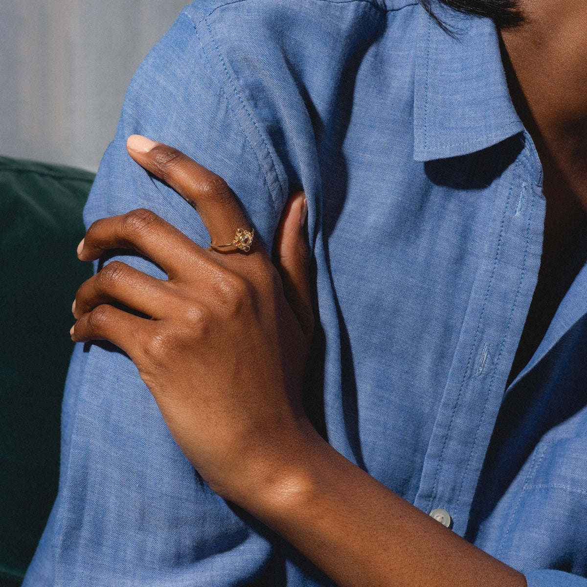 A person in a blue button-up shirt crosses their arms, showcasing the Oberoi Ring—yellow gold plated with tourmaline—on their finger. The image highlights their hands and shirt; part of their face is out of frame.