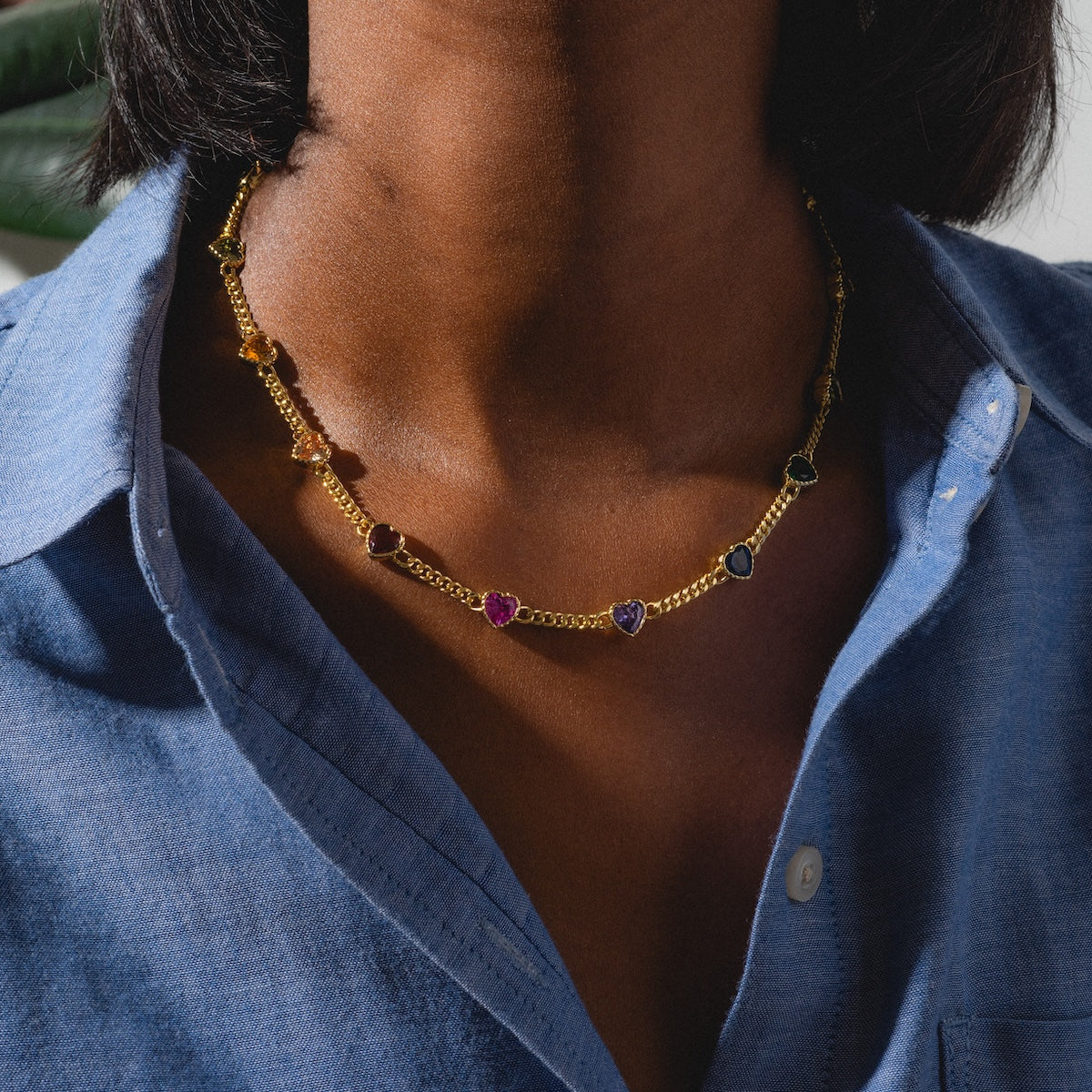 A person wearing a blue button-up shirt and the Love Prism Rainbow Heart Necklace, a 14k gold plated chain adorned with red, purple, yellow, and blue multicolor heart gems.