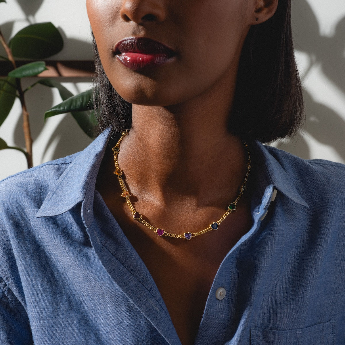 A woman in a blue button-up shirt and dark red lipstick wears the Love Prism Rainbow Heart Necklace, featuring multicolor heart gems, as she stands in natural light framed by plant leaves and shadows.