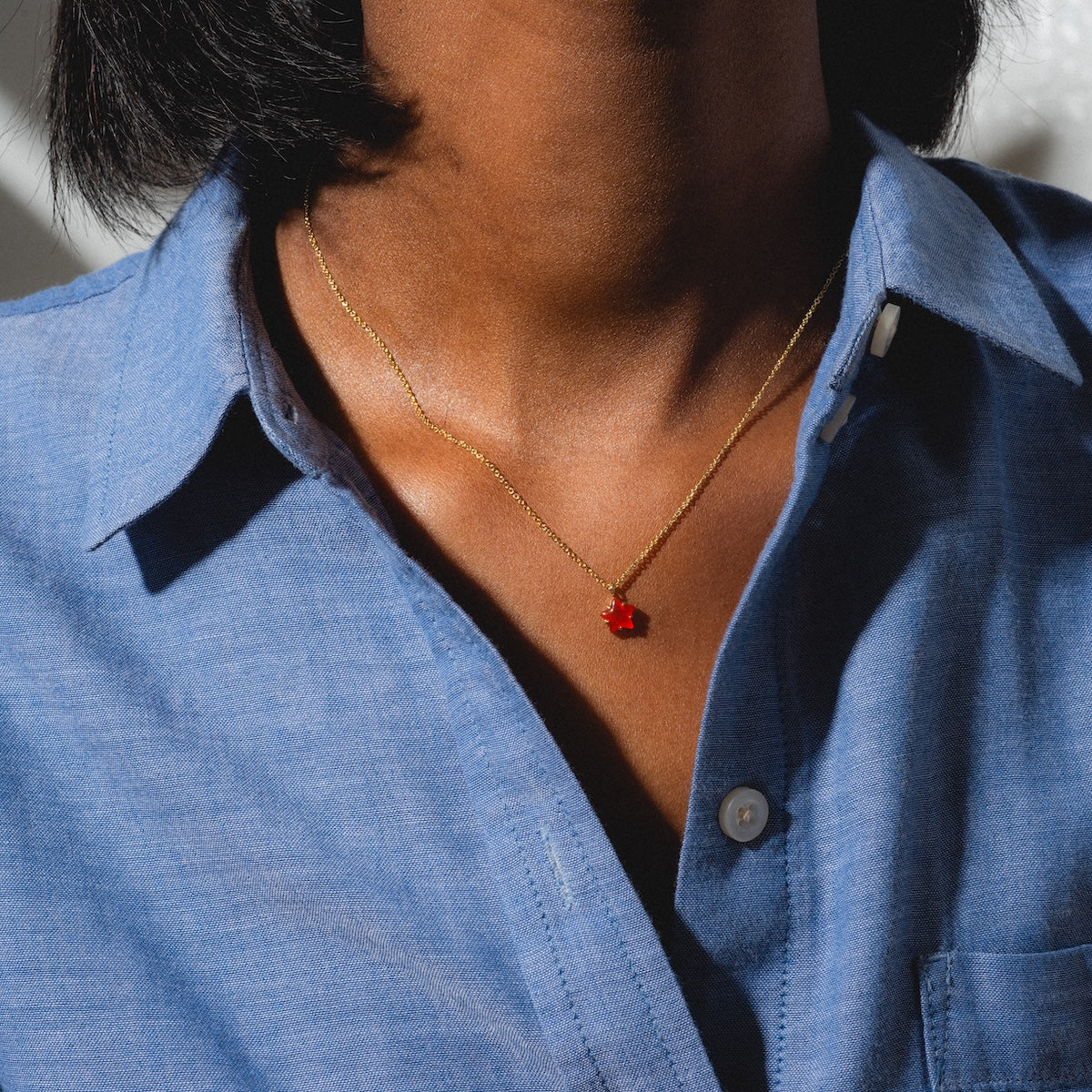 A person wears a blue button-up shirt and the Star Bright Necklace—a gold chain with a small red flower pendant—shown from shoulders to collarbone.