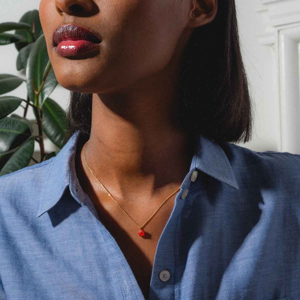 A woman in a light blue shirt with glossy lips wears the Star Bright Necklace, featuring a gold chain and red chalcedony pendant, while standing in bright sunlight beside a leafy plant.