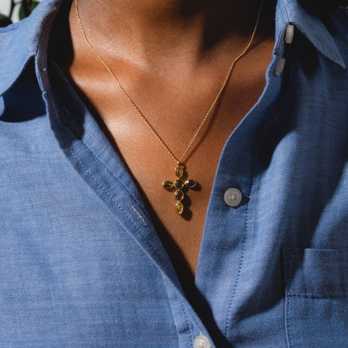 A person in a blue button-up shirt showcases the Anika Chain Necklace, highlighted by a gold cross-shaped pendant with small oval sections, in bright natural light.